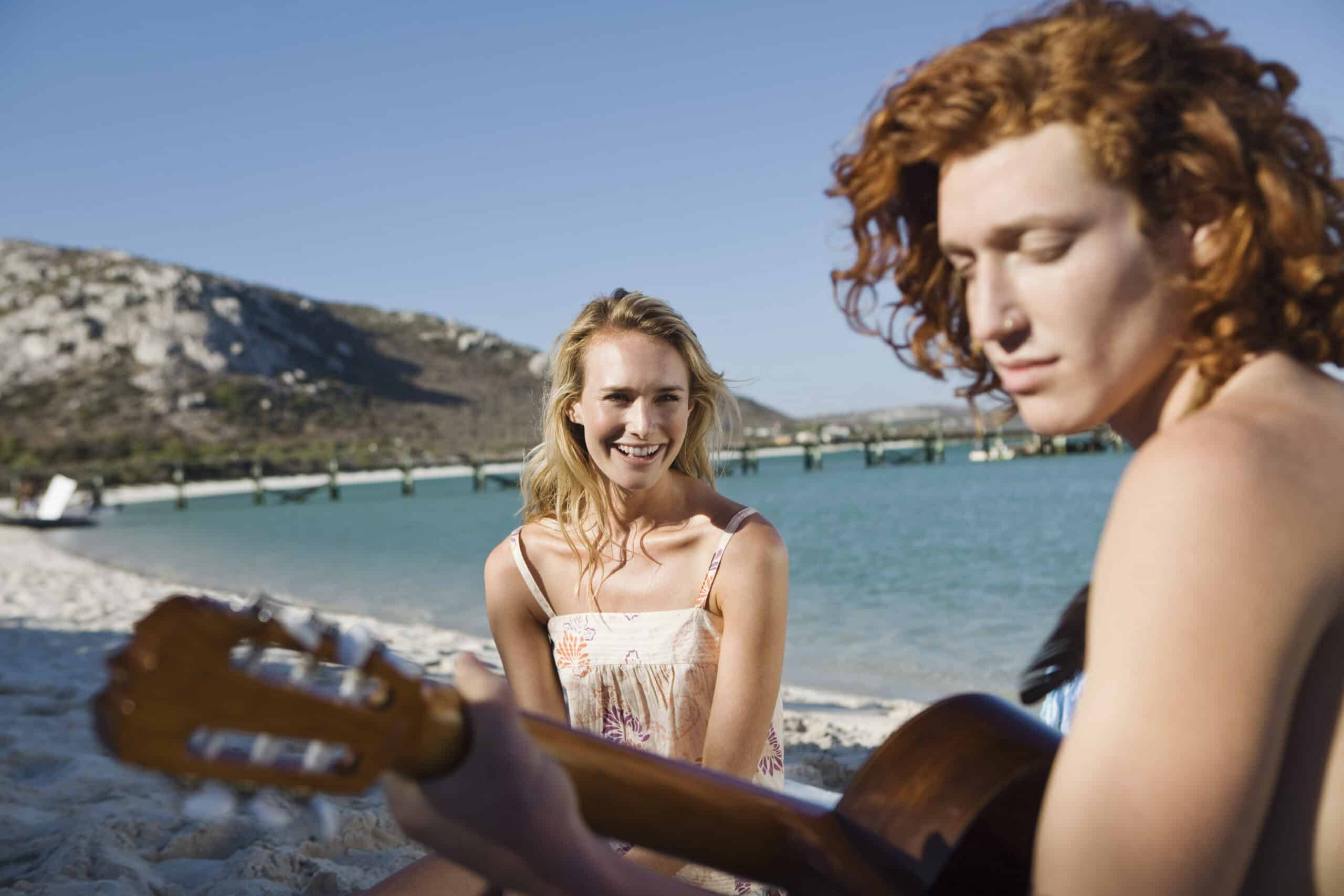 Eine Frau mit roten Haaren spielt Gitarre an einem Sandstrand, während eine andere Frau mit blonden Haaren sie anlächelt, mit Bergen im Hintergrund und Booten im Meer. © Fotografie Tomas Rodriguez