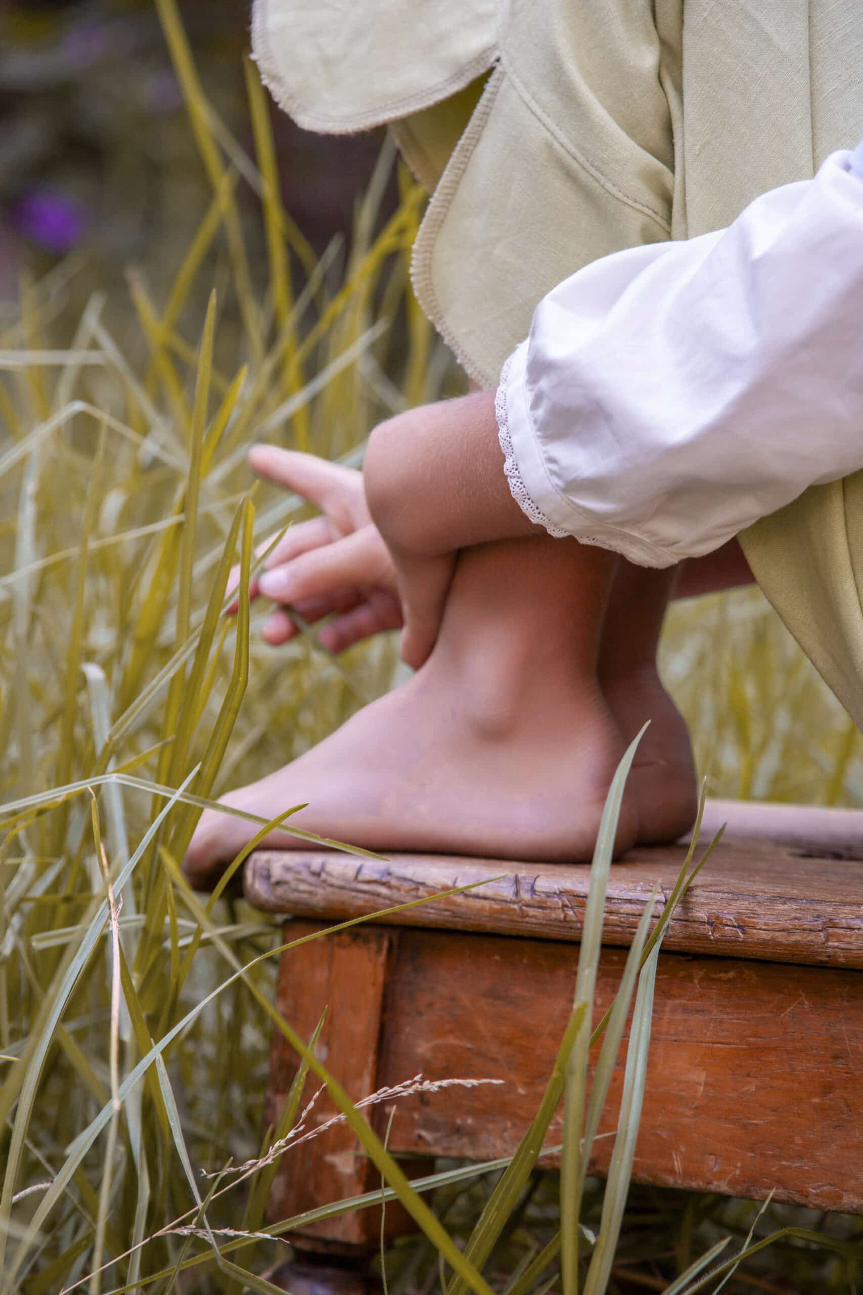 Eine Nahaufnahme des nackten Fußes einer Person, die auf einem Holzhocker im hohen Gras ruht. Sie trägt einen weißen Ärmel mit Spitzendetails, der bis zum Knöchel reicht. © Fotografie Tomas Rodriguez