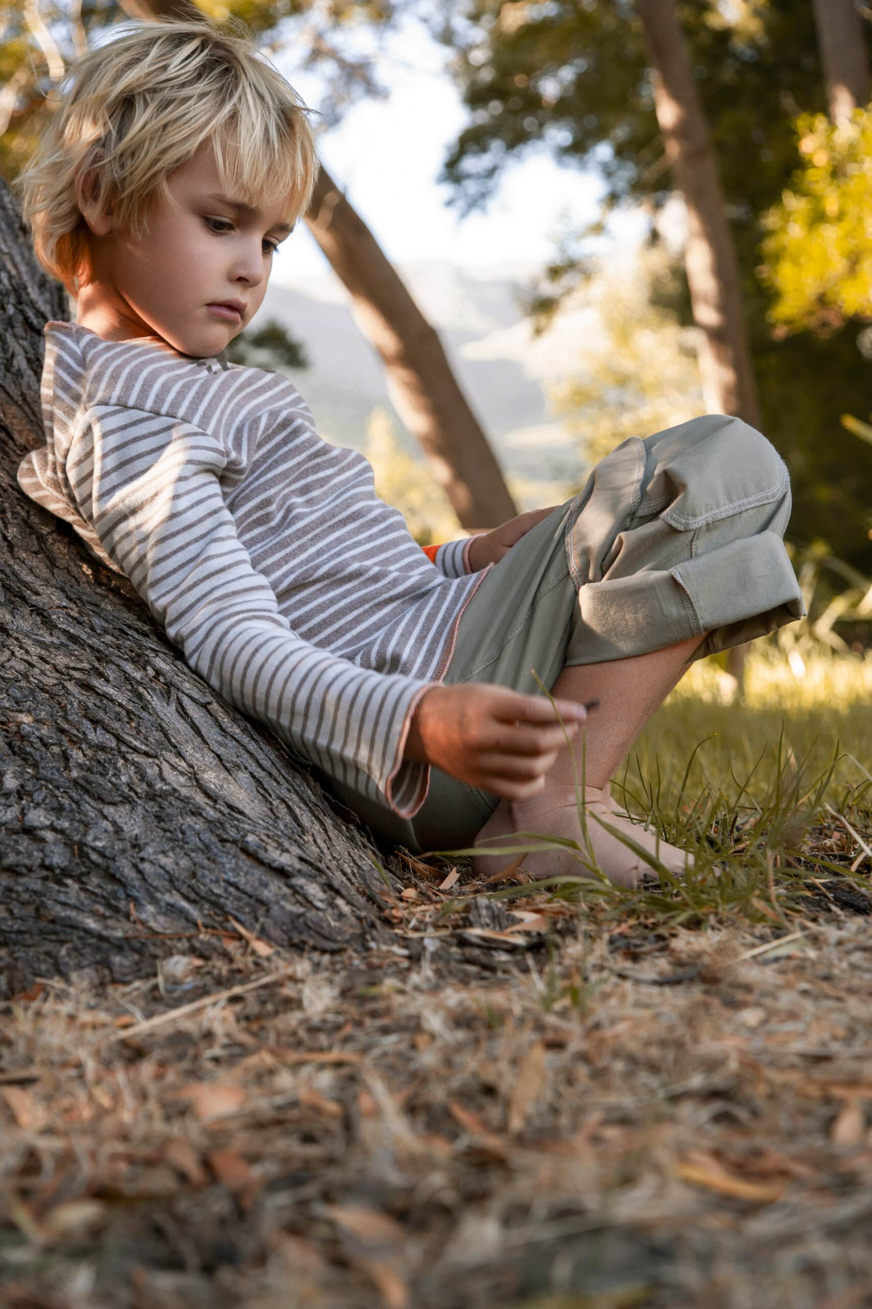 Ein kleiner Junge sitzt im Freien an einen Baumstamm gelehnt und konzentriert sich darauf, seine Schuhe zu binden, umgeben von natürlichem Licht und Laub. Im Hintergrund sind schwach Berge zu erkennen. © Fotografie Tomas Rodriguez