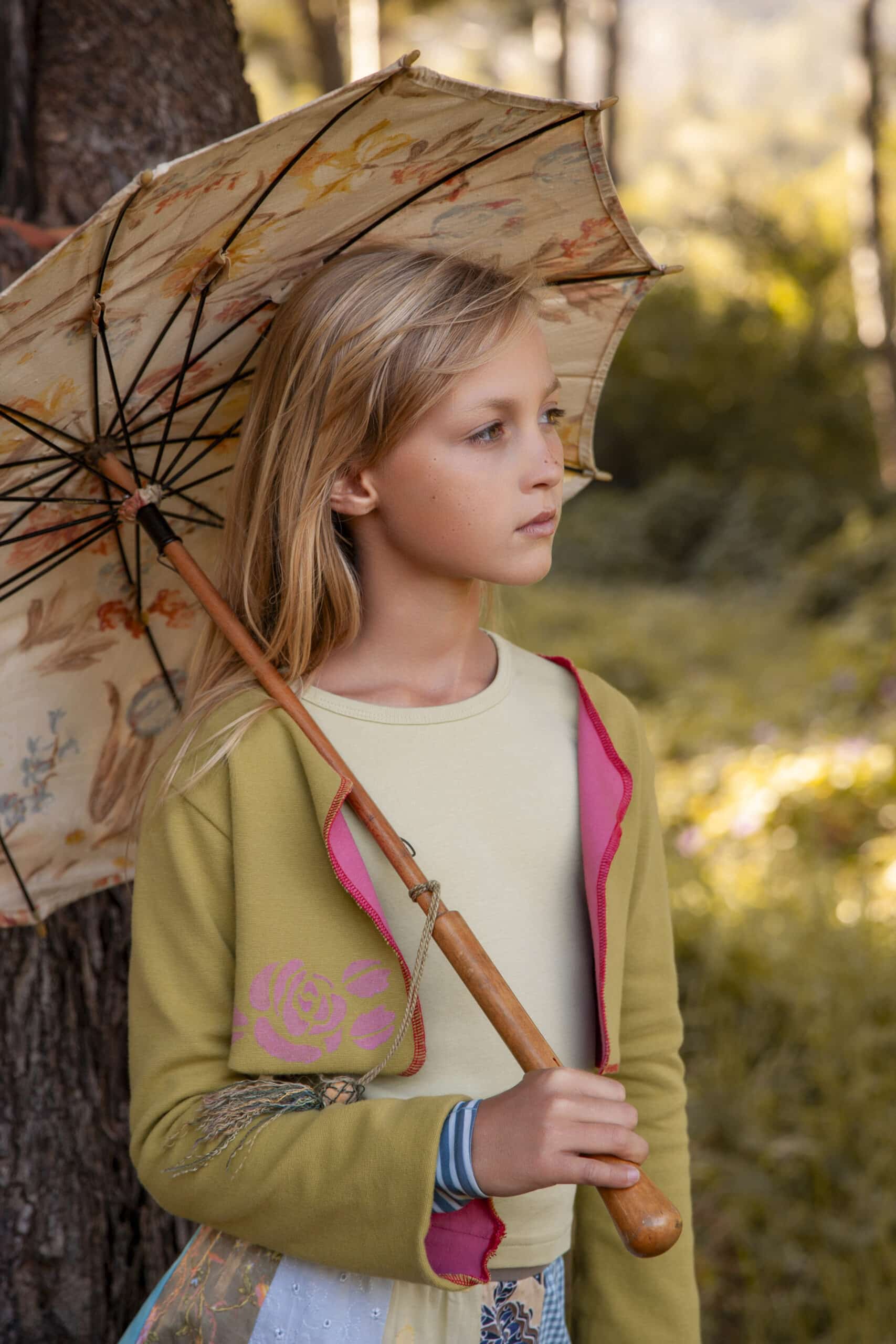 Ein junges Mädchen steht mit einem dekorativen Regenschirm nachdenklich in einem sonnenbeschienenen Wald. Sie trägt einen leichten Pullover und eine kleine braune Tasche, umgeben von Herbstblättern. © Fotografie Tomas Rodriguez