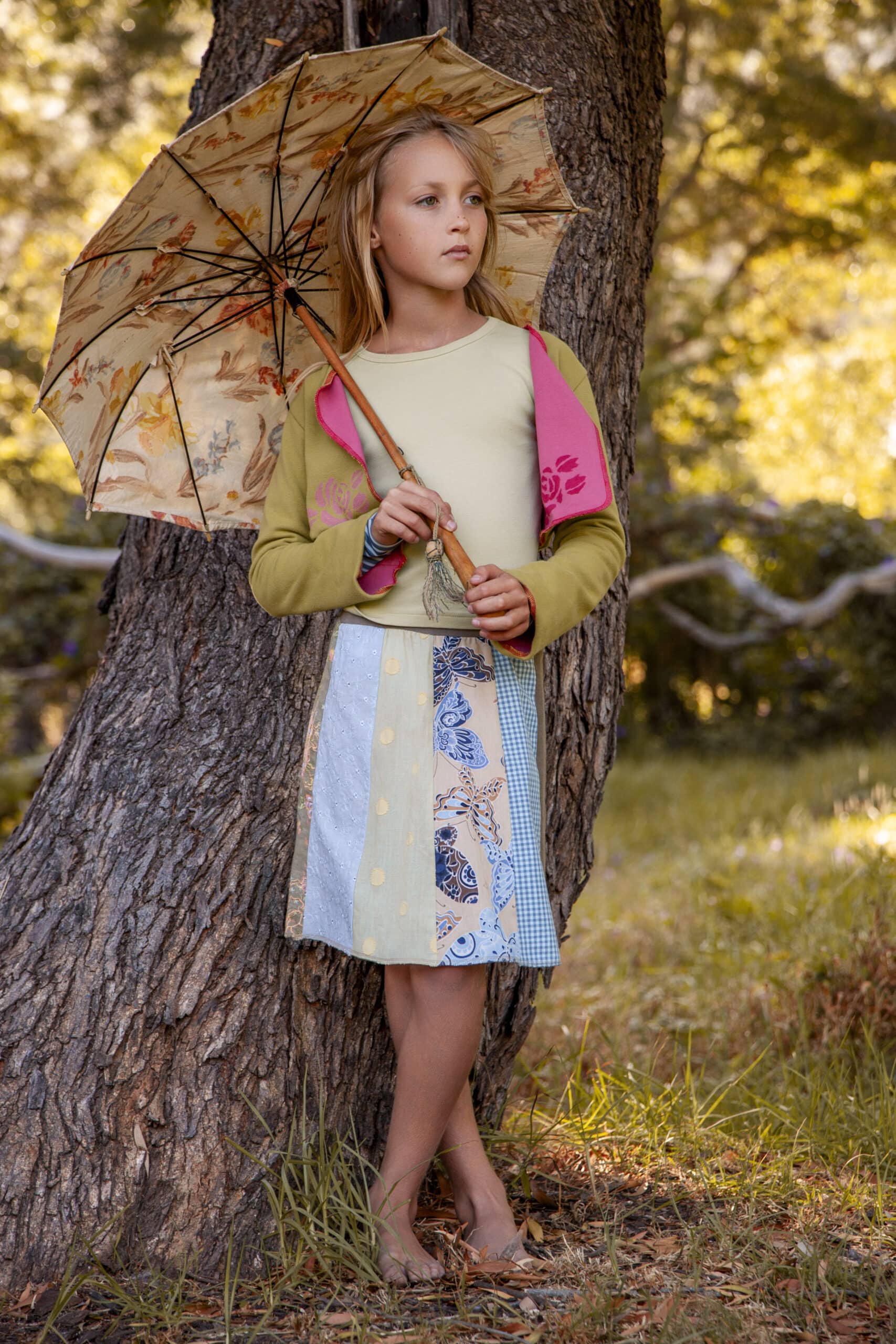 Ein junges Mädchen steht mit einem gemusterten Regenschirm in der Hand unter einem Baum, trägt einen bunten Patchworkrock und einen pastellfarbenen Pullover und blickt nachdenklich in die Ferne. © Fotografie Tomas Rodriguez