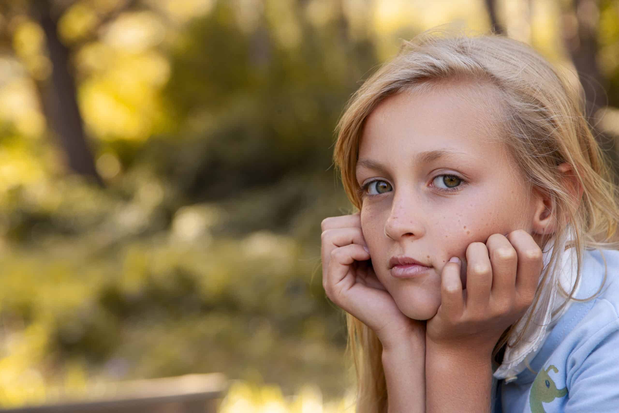 Ein nachdenkliches junges Mädchen mit konzentriertem Gesichtsausdruck stützt ihr Kinn auf ihre Hände, vor einem leicht verschwommenen Hintergrund aus grünem Laub. © Fotografie Tomas Rodriguez