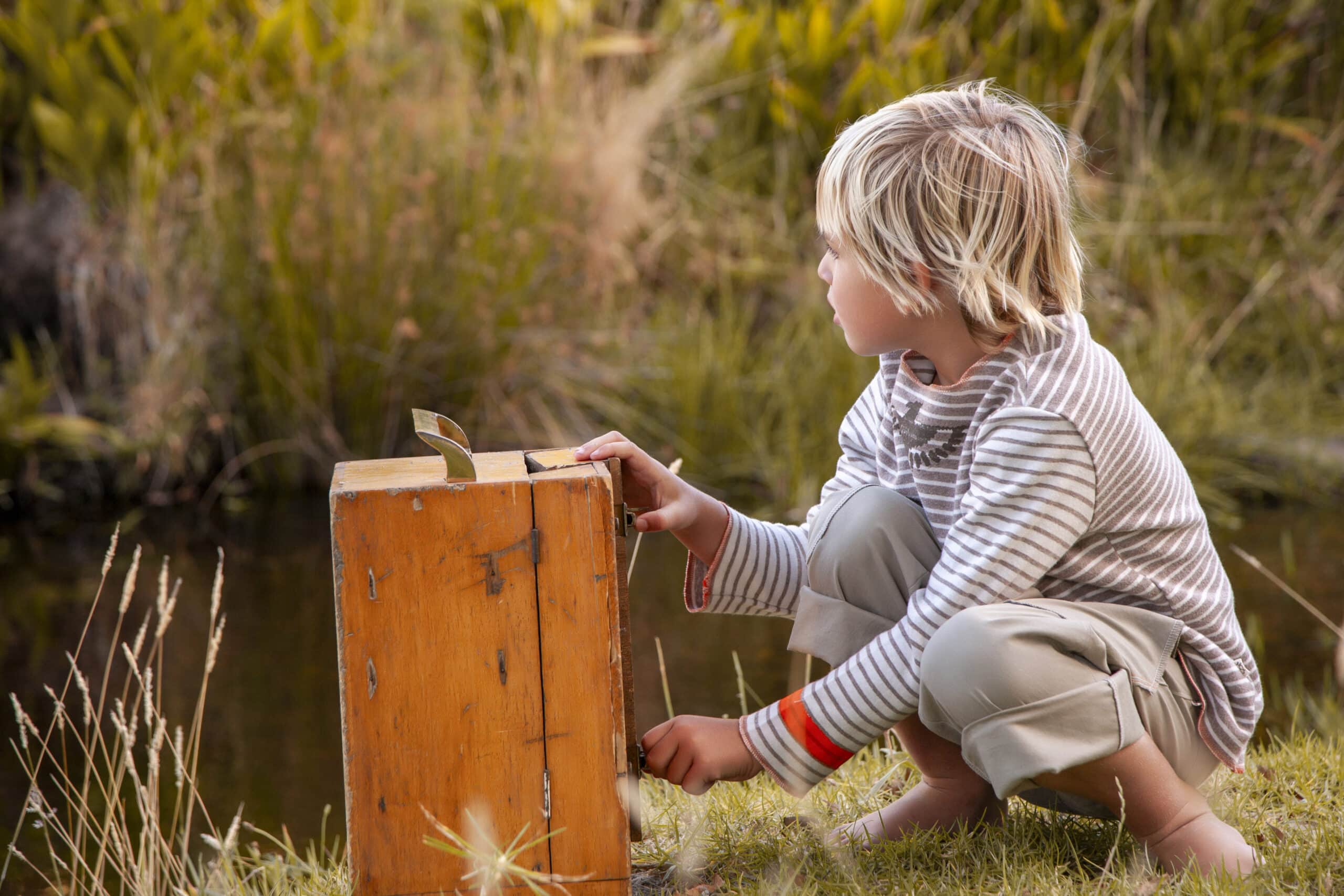 Ein kleines Kind mit blondem Haar, das ein gestreiftes Langarmshirt und Khaki-Shorts trägt, kniet neben einem Teich und beobachtet aufmerksam eine Holzkiste mit einem Buch darin in einer natürlichen Umgebung. © Fotografie Tomas Rodriguez