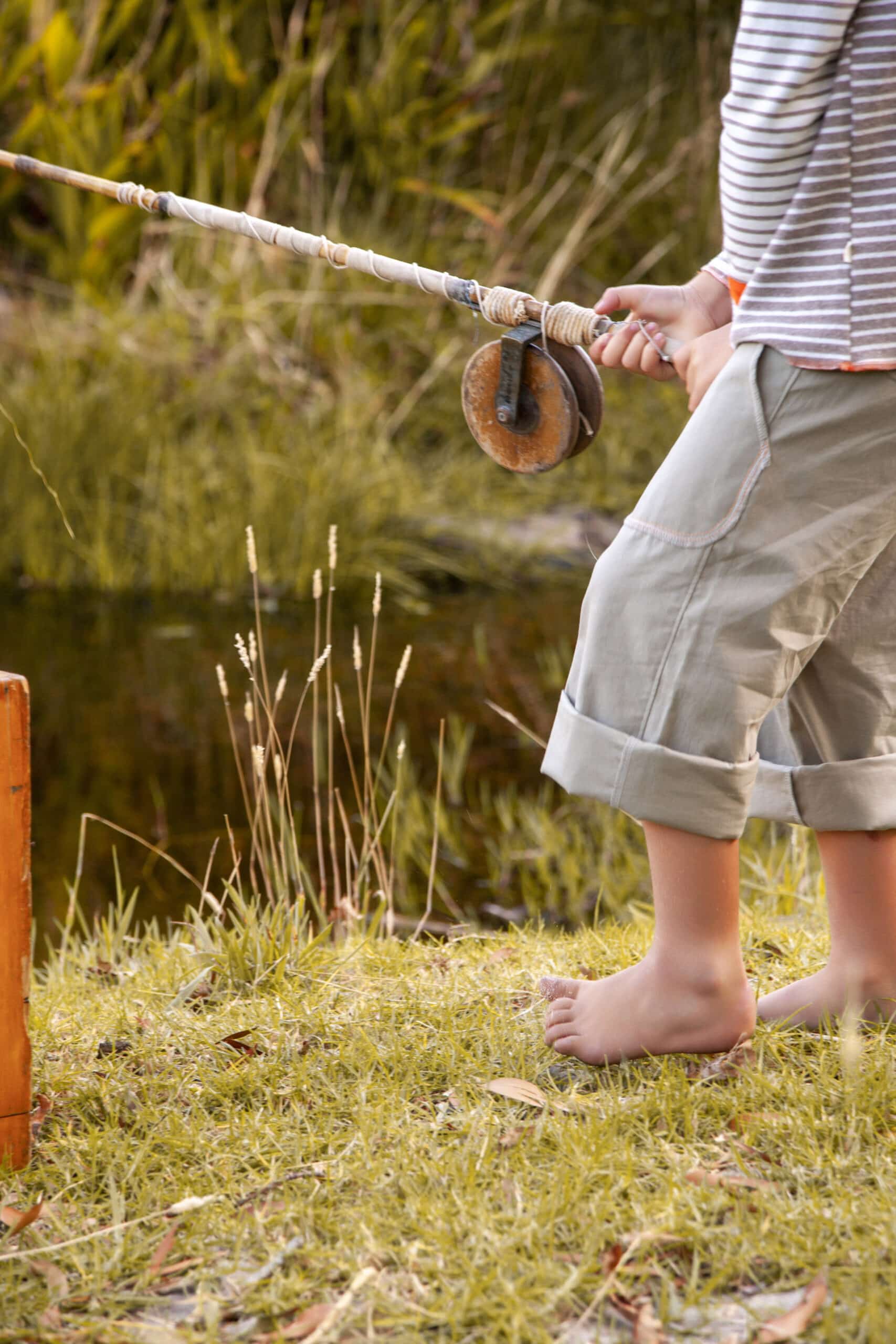 Ein Kind steht barfuß im Gras neben einem Teich und hält eine improvisierte Angelrute aus einem Stock und einer Rolle in der Hand. Der Fokus des Kindes scheint auf dem Wasser zu liegen, was auf einen Moment hoffnungsvoller Erwartung hindeutet. © Fotografie Tomas Rodriguez