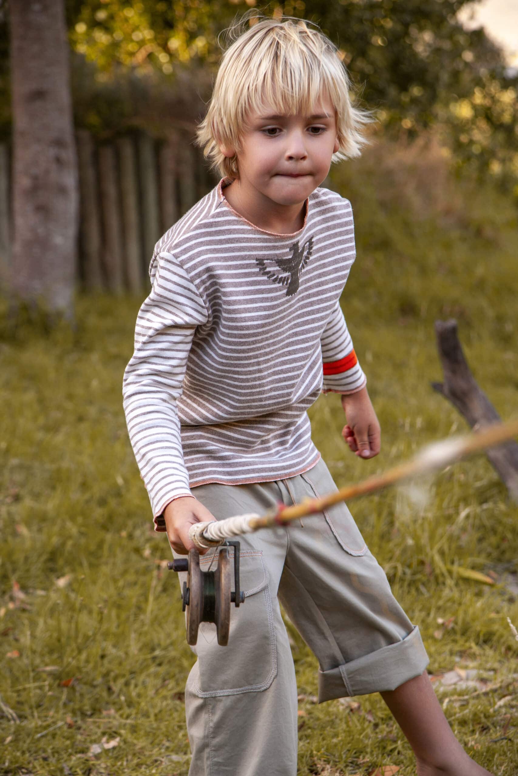 Ein kleiner Junge mit blondem Haar spielt im Freien. Er zieht konzentriert an einem Seil, das an einem hölzernen Flaschenzug befestigt ist. Er trägt ein gestreiftes Langarmshirt und Shorts und steht auf einer Rasenfläche mit Bäumen im Hintergrund. © Fotografie Tomas Rodriguez