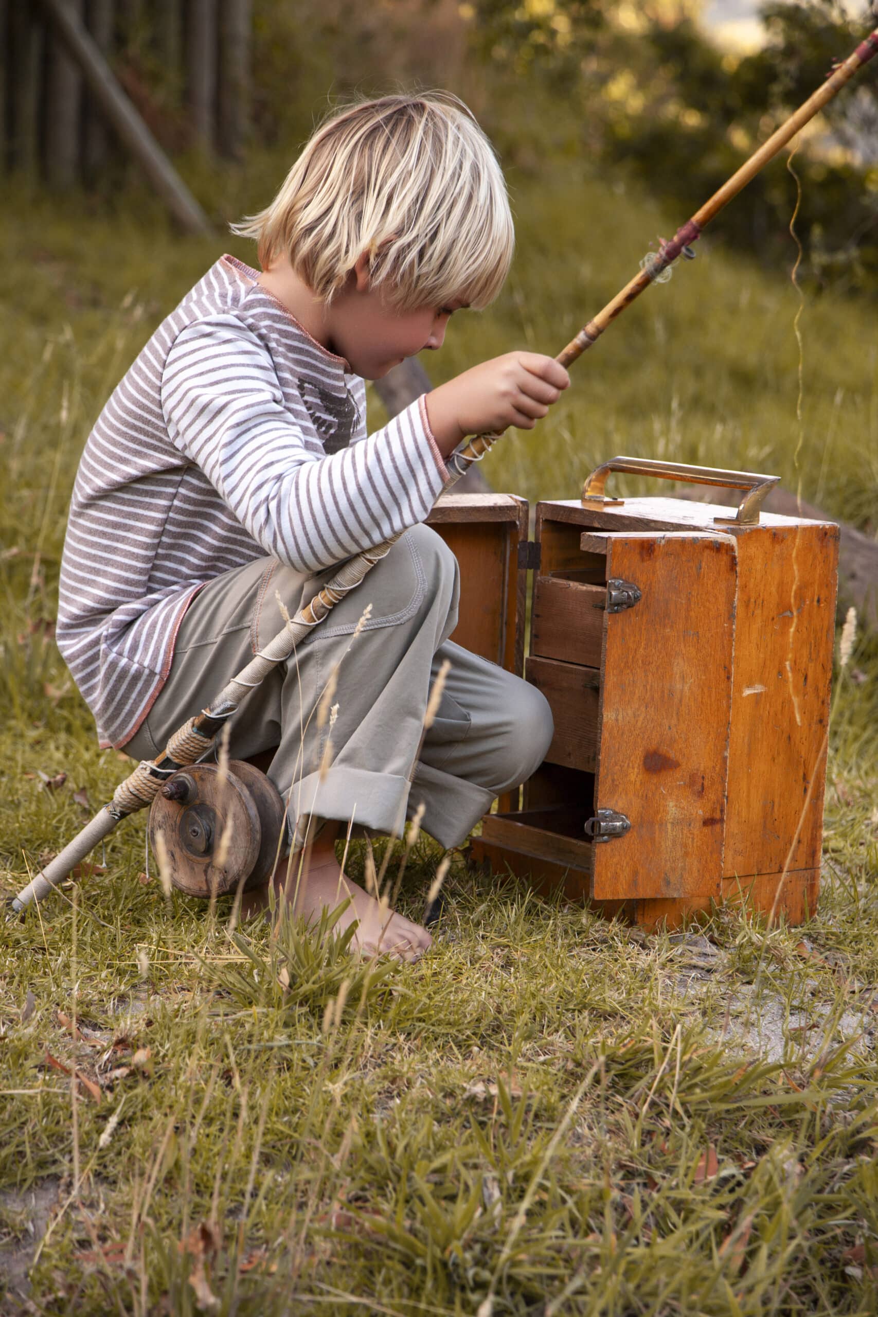 Ein kleiner Junge mit blondem Haar sitzt im Gras und konzentriert sich ganz aufs Angeln. Er verwendet eine improvisierte Angelrute mit Rolle und hat neben sich eine Blechdose und eine Spule. © Fotografie Tomas Rodriguez