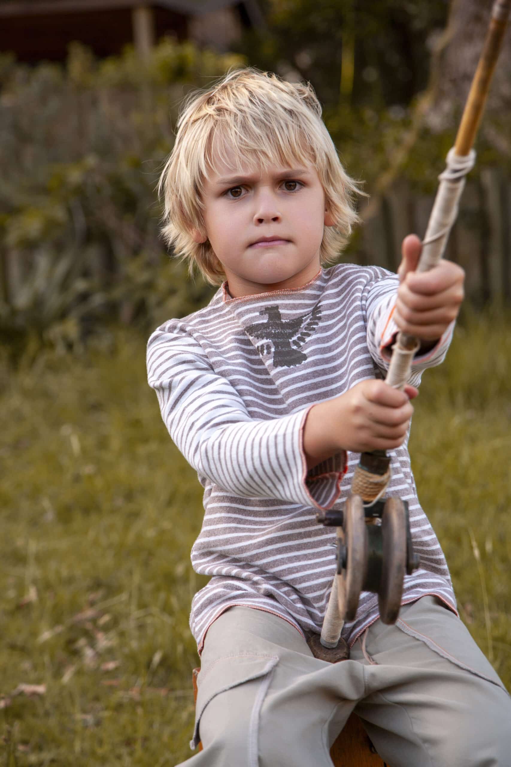 Ein kleiner Junge mit zerzaustem blondem Haar sitzt mit konzentriertem Gesichtsausdruck auf einer Seilschaukel, hält sich fest am Seil fest. Er ist im Freien, umgeben von Grün. © Fotografie Tomas Rodriguez