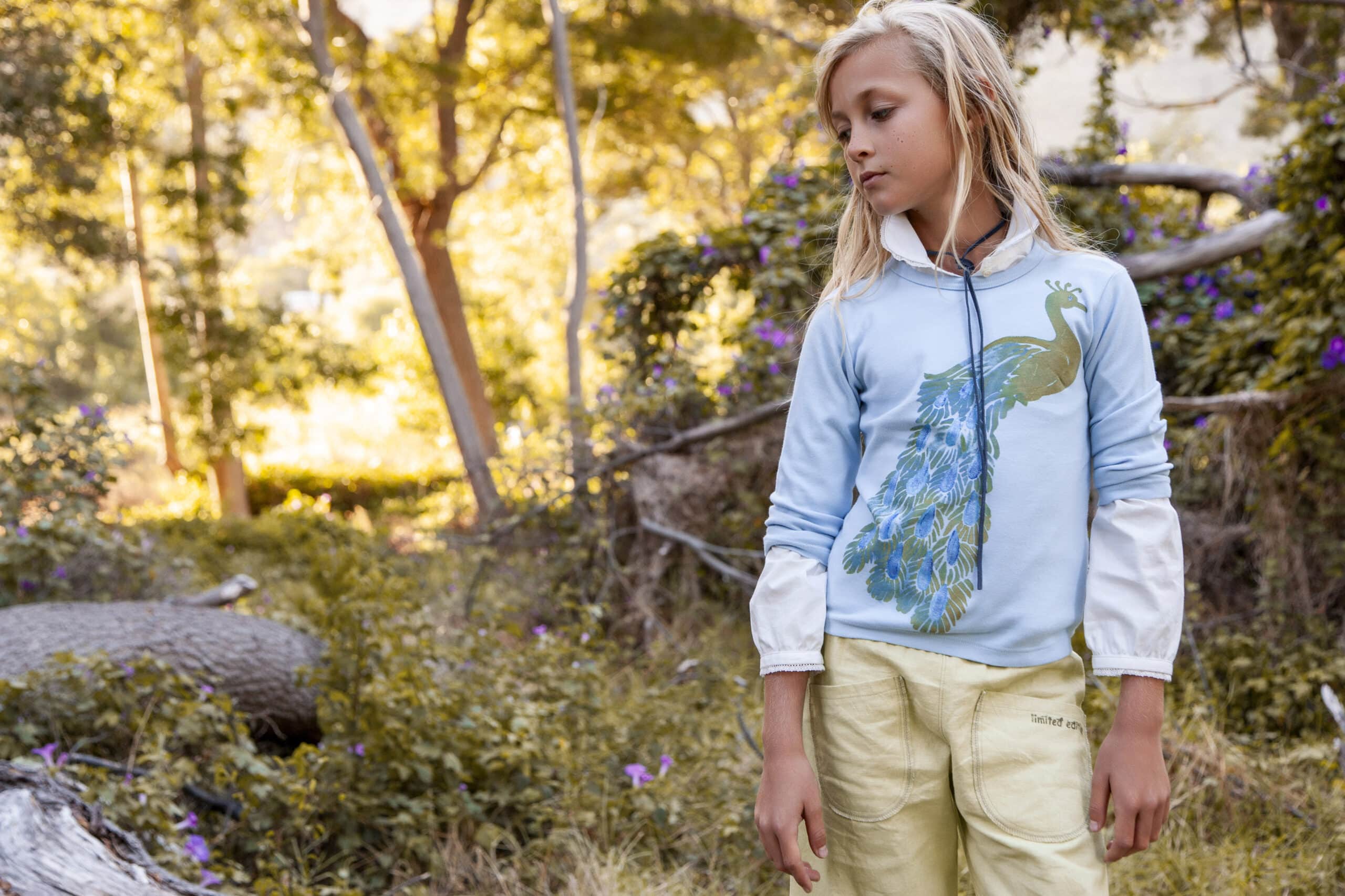 Ein junges Mädchen in einem hellblauen Kapuzenpullover und gelben Hosen geht nachdenklich durch einen sonnendurchfluteten Garten mit üppigem Grün und lila Blumen. © Fotografie Tomas Rodriguez