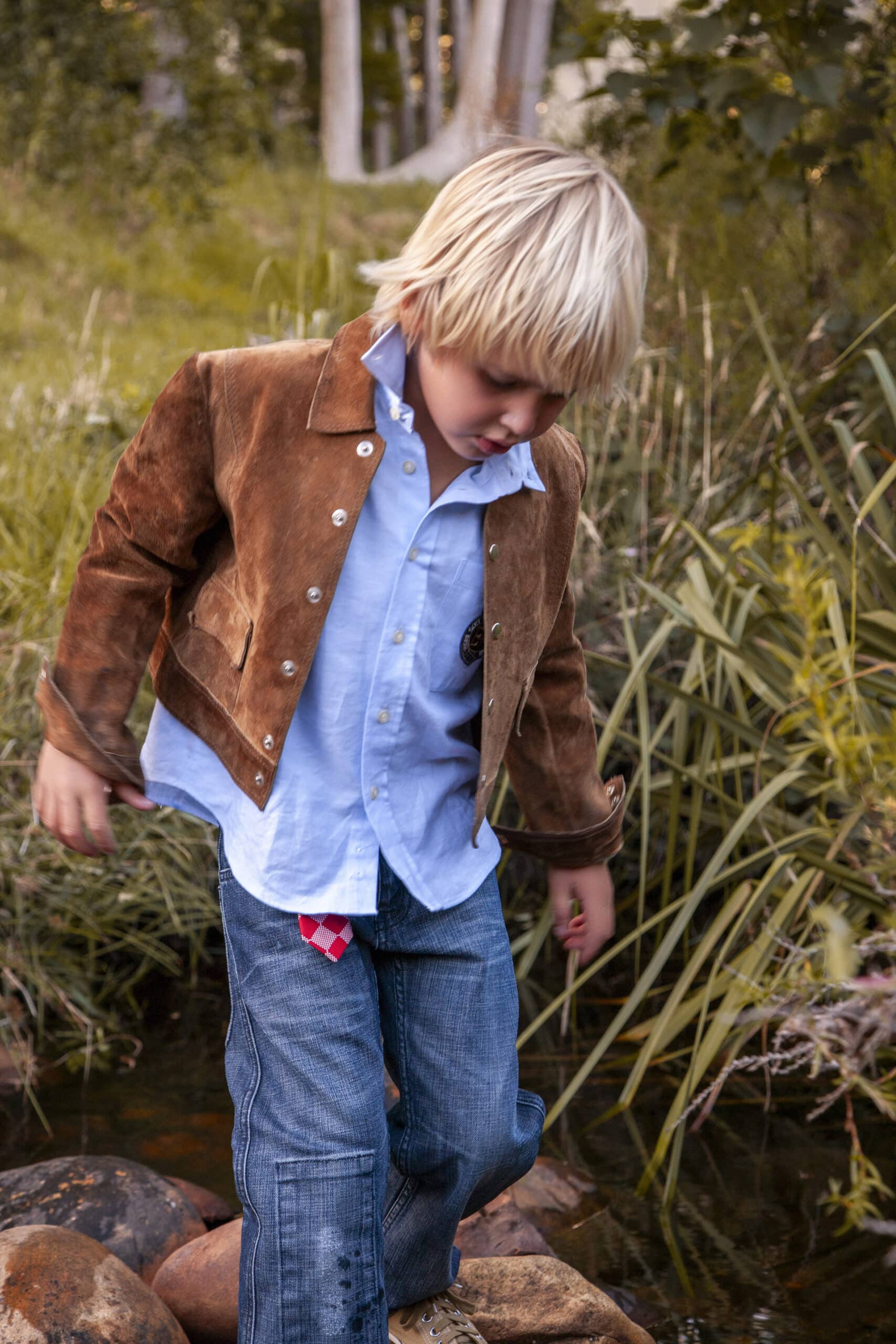 Ein kleiner Junge mit blondem Haar, einer braunen Wildlederjacke, einem blauen Hemd und Jeans, tritt vorsichtig auf Steine neben einem Bach, der von Gras und Schilf umgeben ist. © Fotografie Tomas Rodriguez