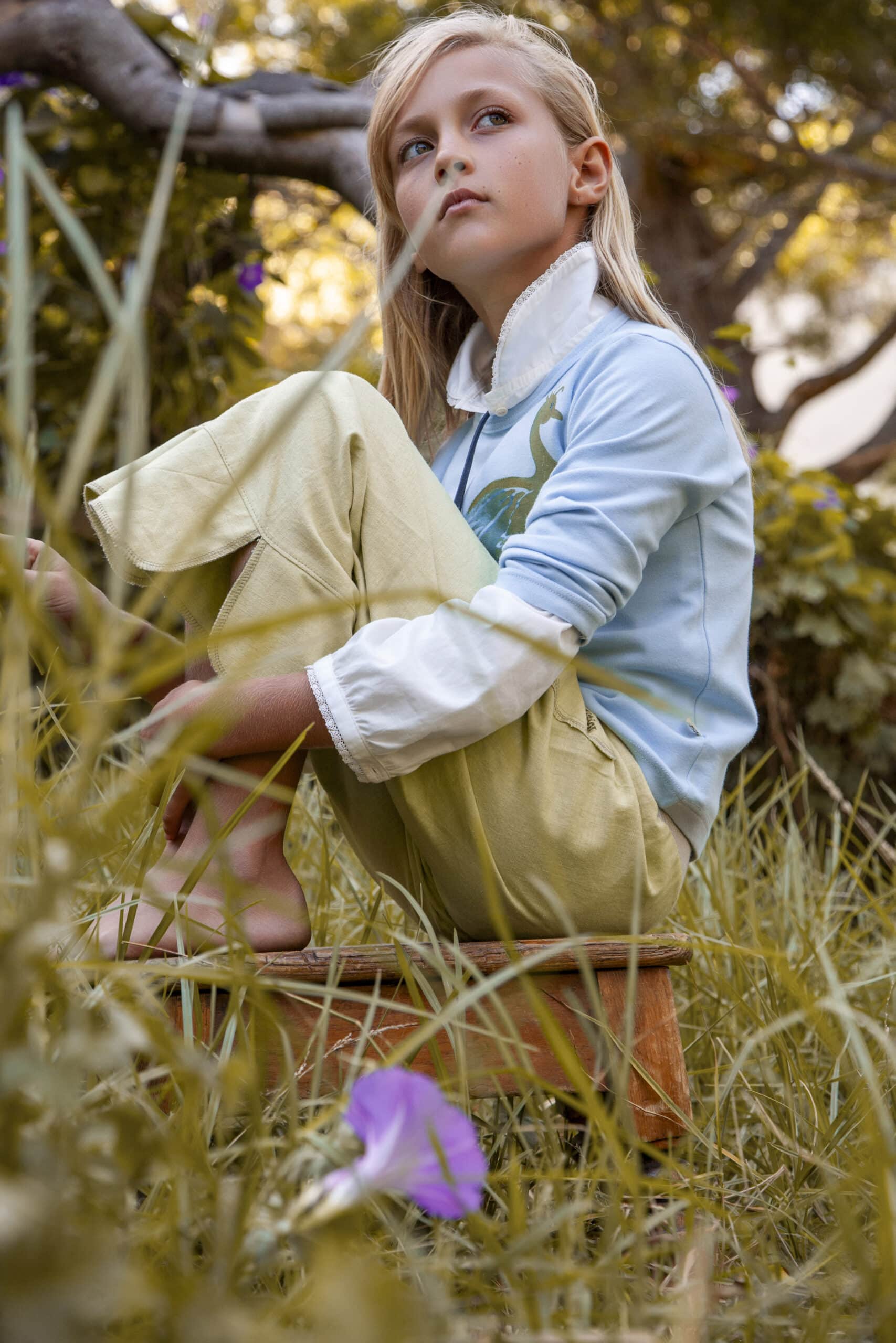 Ein junges Mädchen mit blondem Haar und einem blau-gelben Outfit sitzt nachdenklich unter einem Baum in einem Garten, mit sanftem Fokus auf das umgebende Grün und eine violette Blume. © Fotografie Tomas Rodriguez