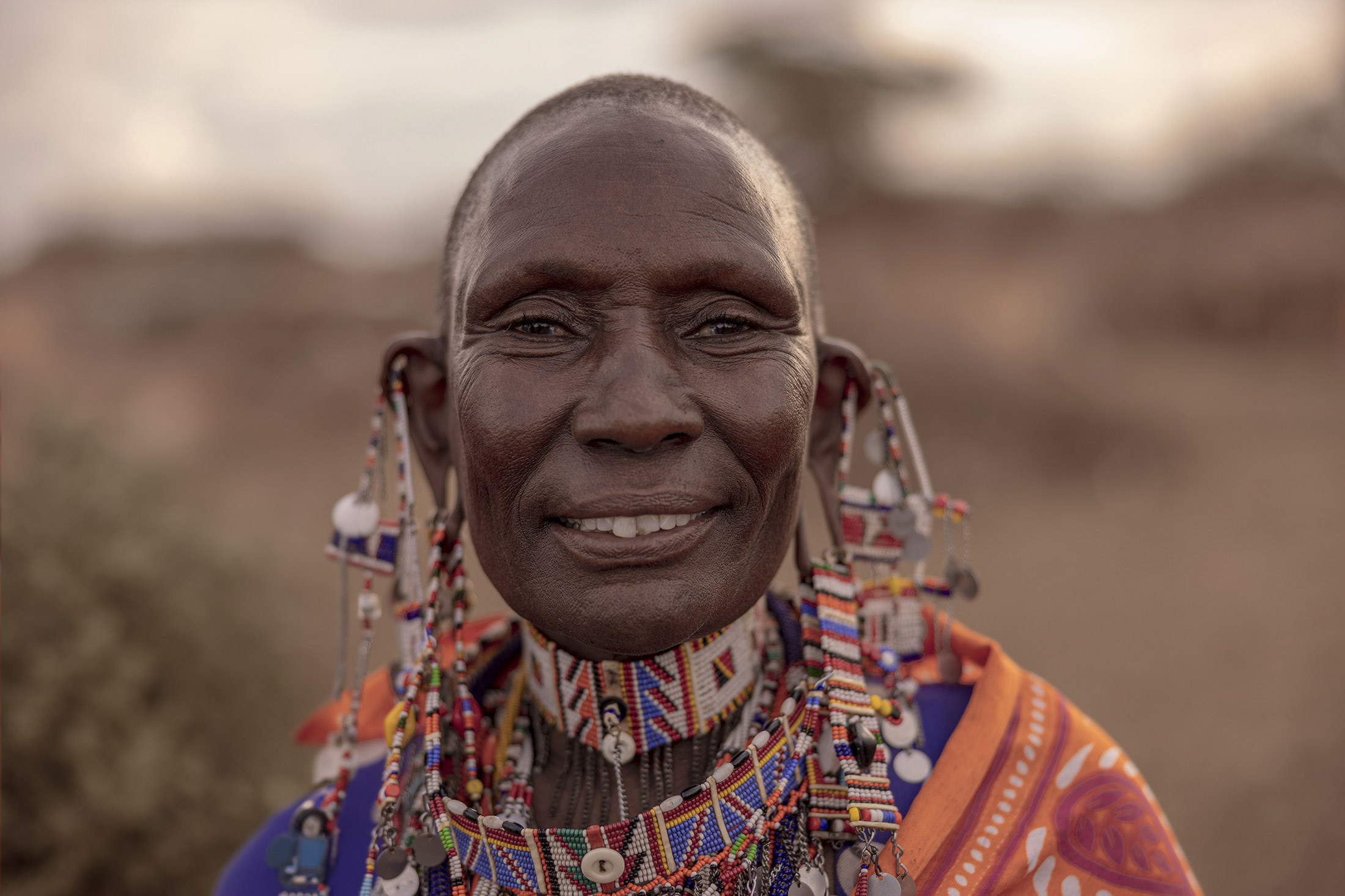 Ein Nahaufnahme-Porträt einer älteren Massai-Frau, geschmückt mit bunten Perlenketten und Ohrringen, in traditioneller orangefarbener Kleidung gekleidet, sanft lächelt vor einem unscharfen natürlichen Hintergrund. © Fotografie Tomas Rodriguez