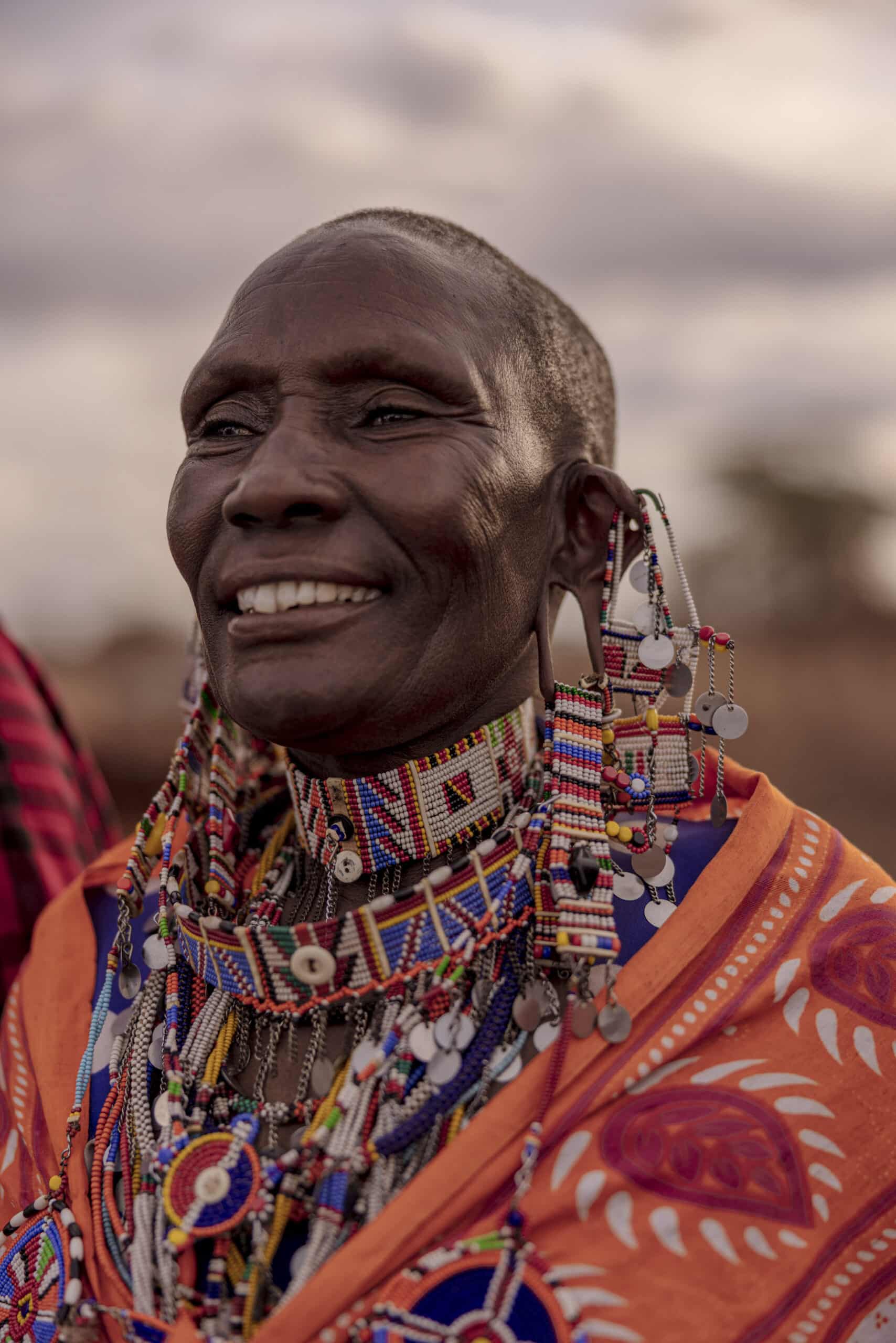 Eine ältere Massai-Frau in traditioneller Kleidung, geschmückt mit bunten Perlenketten und Ohrringen, lächelt breit im Freien unter einem sanften, dämmrigen Himmel. © Fotografie Tomas Rodriguez