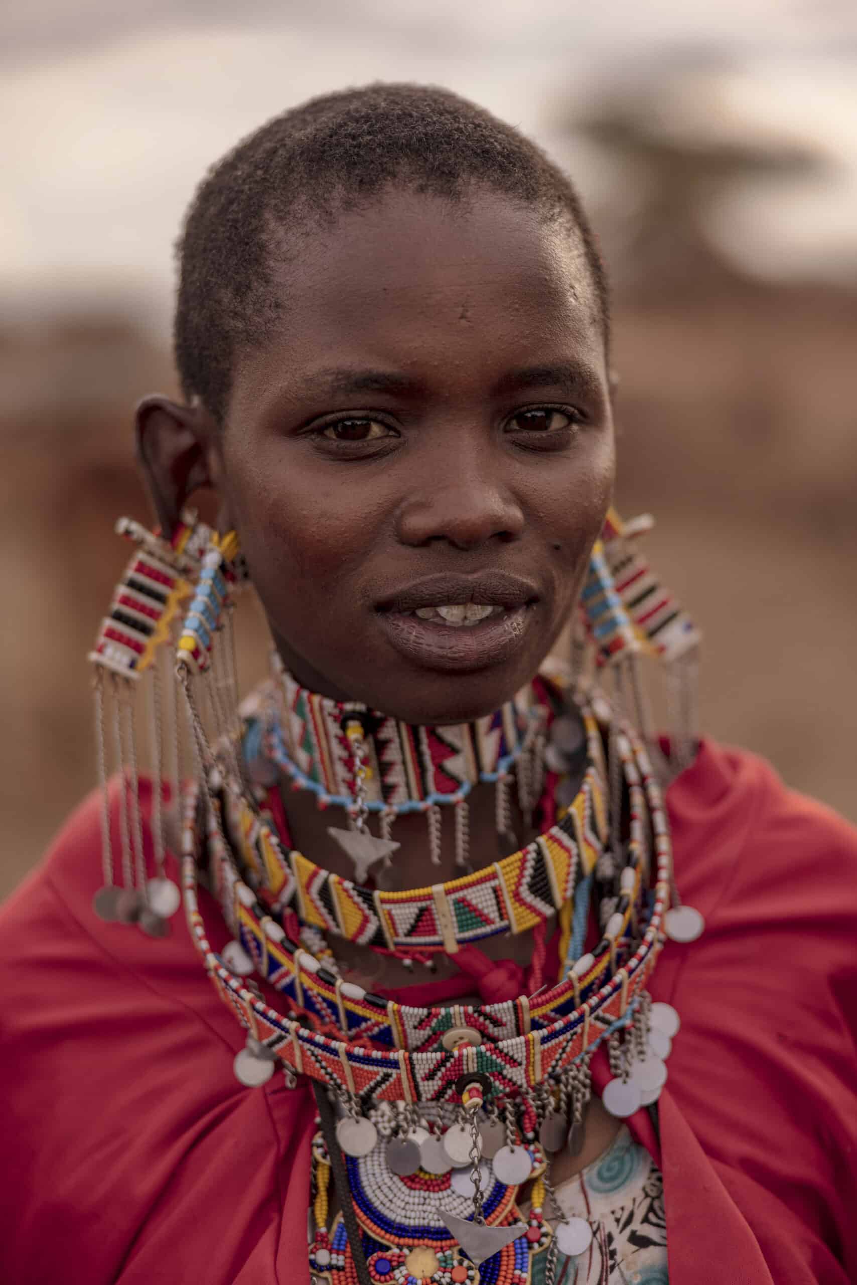 Eine Massai-Frau in traditioneller Kleidung, mit einem leuchtend roten Schal und kunstvollen Perlenketten, steht vor einem unscharfen Hintergrund. Ihr Gesichtsausdruck ist ruhig und direkt. © Fotografie Tomas Rodriguez