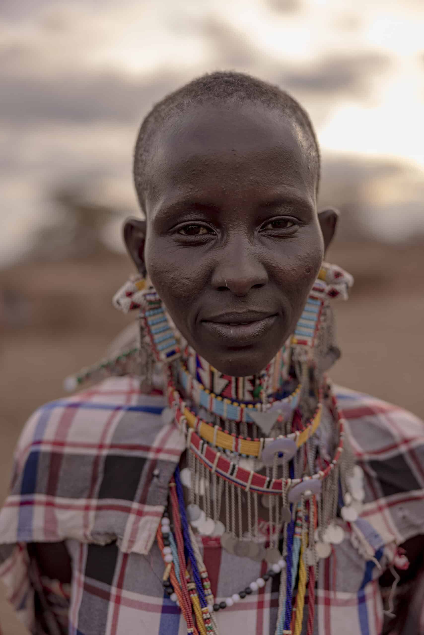 Ein Porträt einer Massai-Frau, geschmückt mit traditionellen Perlenketten und Ohrringen, eine gemusterte Shuka tragend, vor einem weichgezeichneten Wüstenhintergrund. © Fotografie Tomas Rodriguez