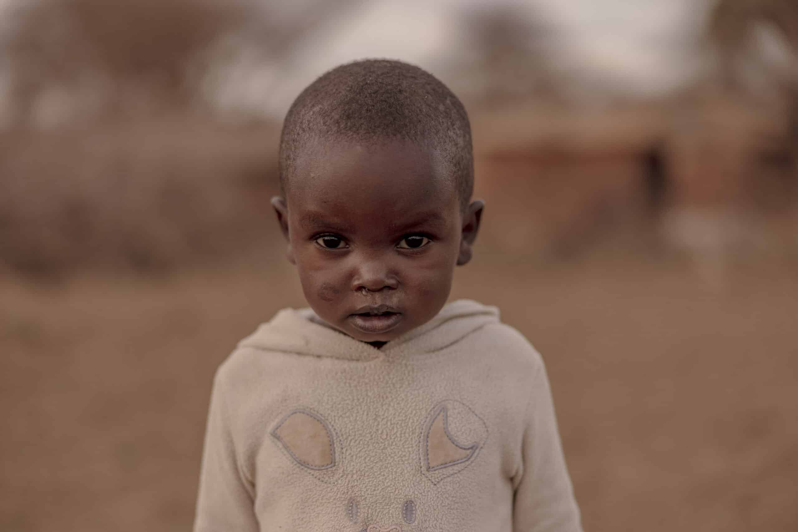 Ein kleines Kind mit ernster Miene und einem abgetragenen Sweatshirt steht in einer staubigen Umgebung mit unscharfem Hintergrund. © Fotografie Tomas Rodriguez