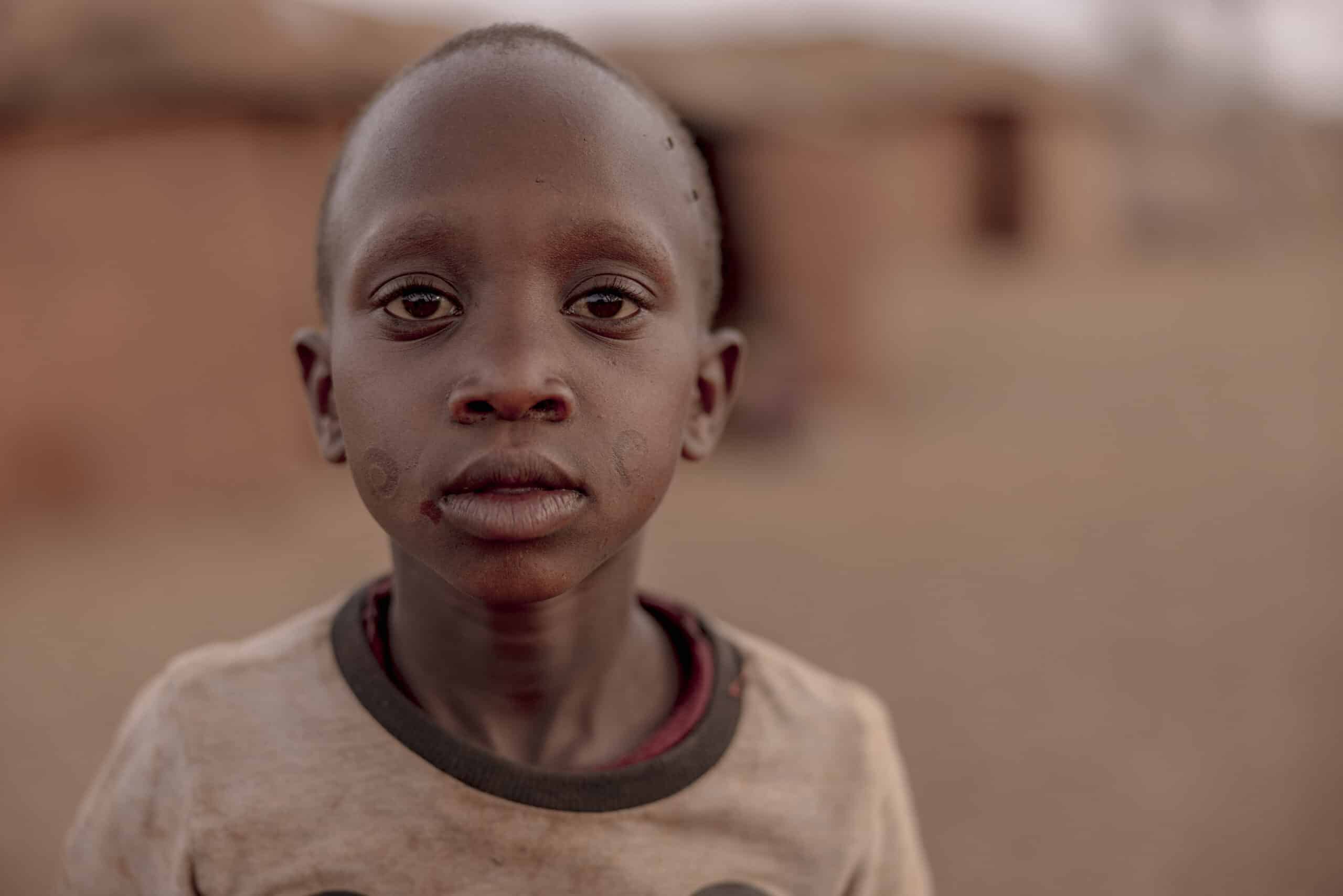 Eine Nahaufnahme eines jungen Jungen mit durchdringendem Blick, der ein abgetragenes T-Shirt trägt und vor dem unscharfen Hintergrund einer sandigen, erdfarbenen Umgebung steht. © Fotografie Tomas Rodriguez