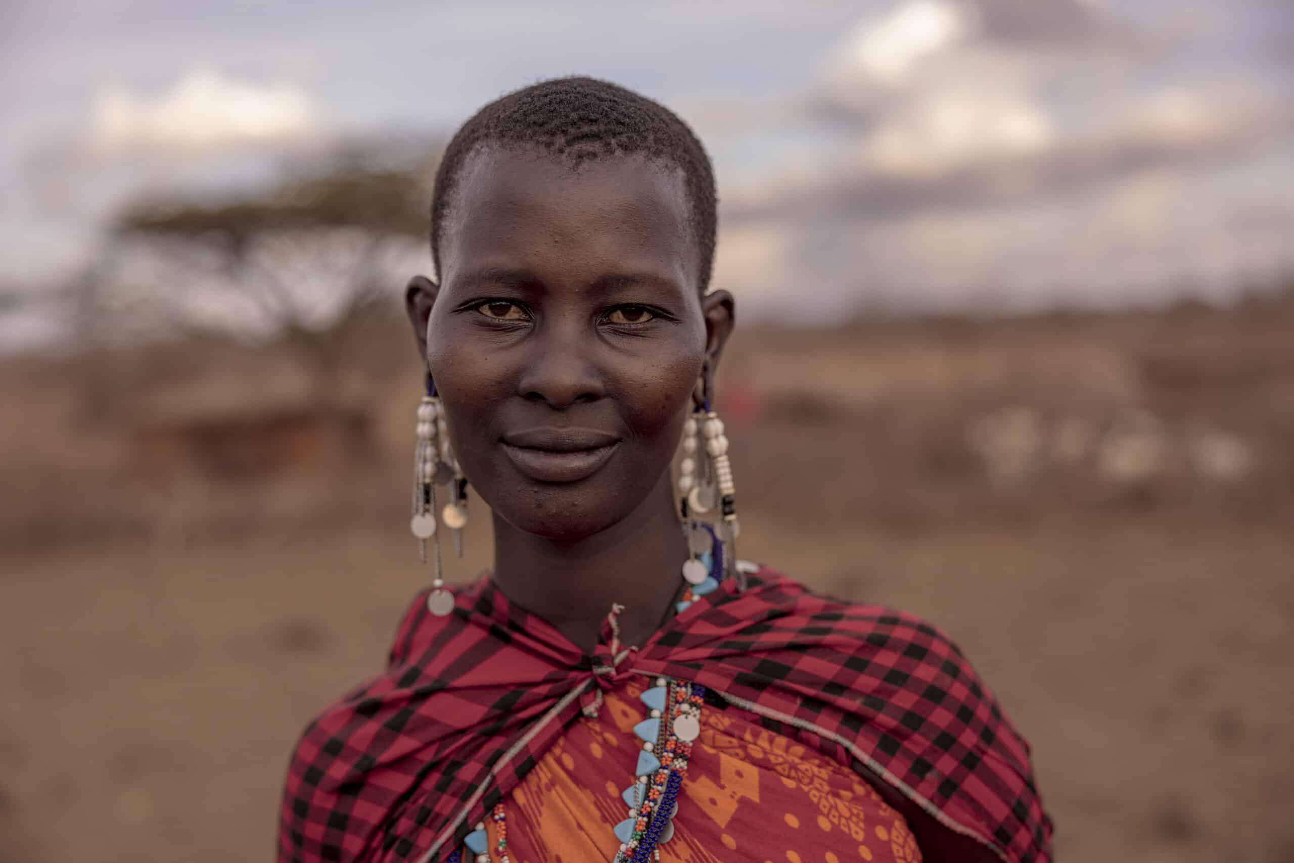 Ein Porträt einer jungen Frau in traditioneller Massai-Kleidung, darunter Perlenohrringe und ein leuchtend rotes Shuka. Sie steht mit heiterem Gesichtsausdruck im Freien vor einer leicht verschwommenen Savannenkulisse. © Fotografie Tomas Rodriguez