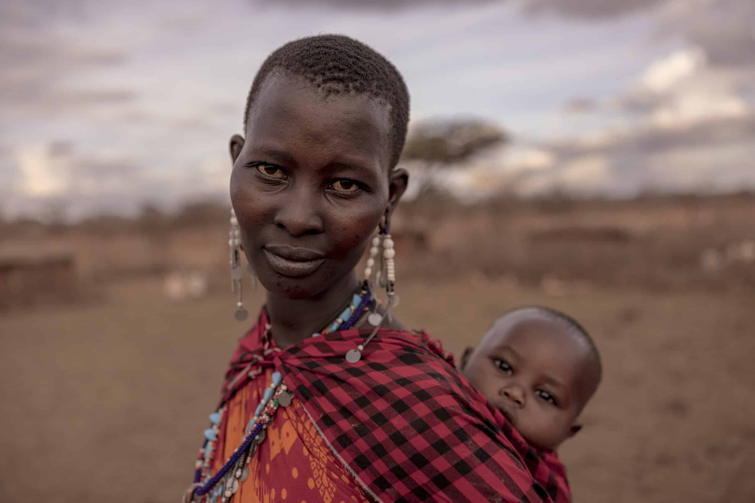 Eine Frau in traditioneller Kleidung mit kunstvollen Ohrringen steht in einer trockenen Landschaft und hält ein Baby, das über ihre Schulter schaut. Beide blicken in die Kamera. © Fotografie Tomas Rodriguez