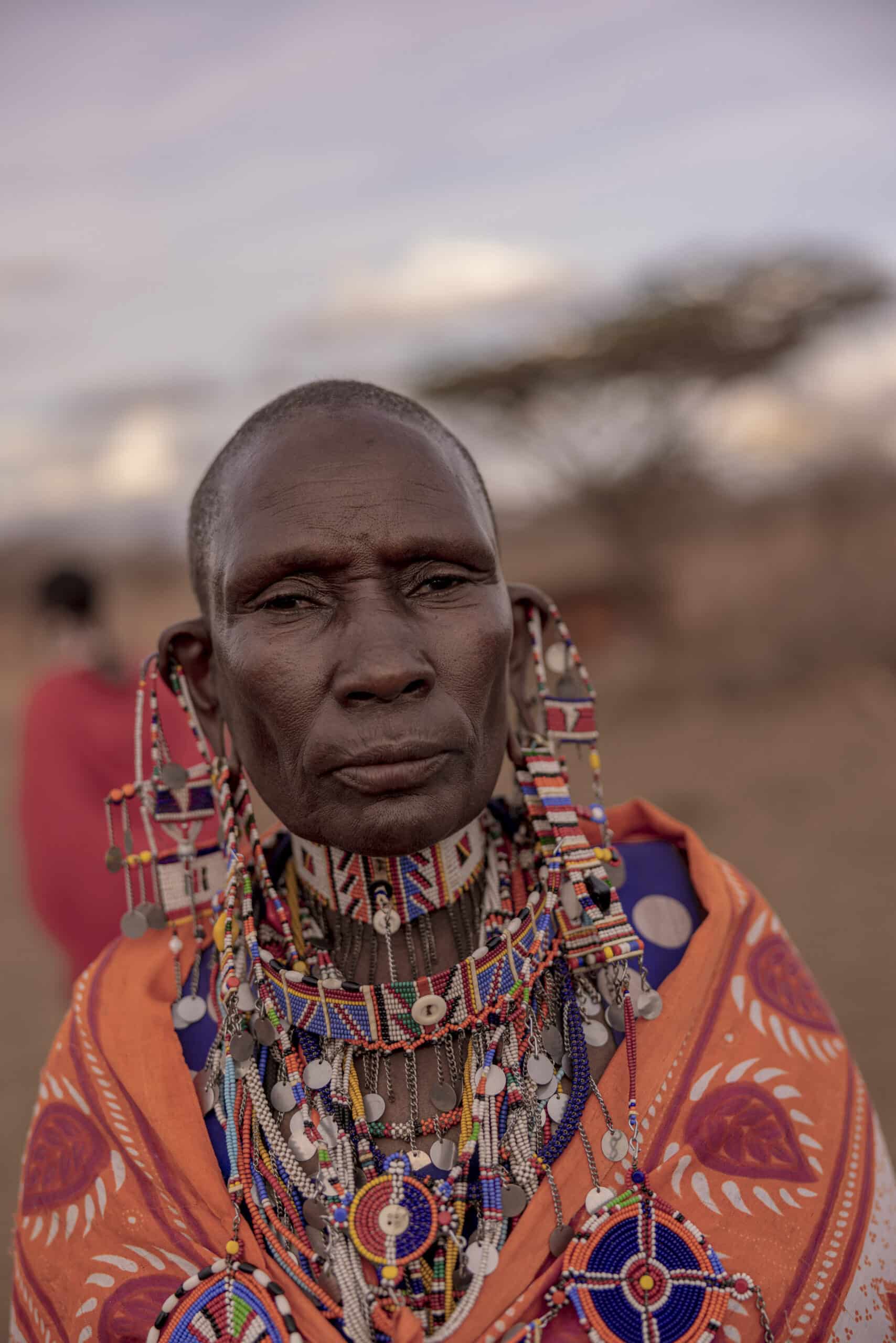 Porträt einer Massai-Frau, geschmückt mit traditionellem Schmuck und leuchtend orangefarbener Kleidung, die in einer halbtrockenen Landschaft mit unscharfem Hintergrund steht. © Fotografie Tomas Rodriguez