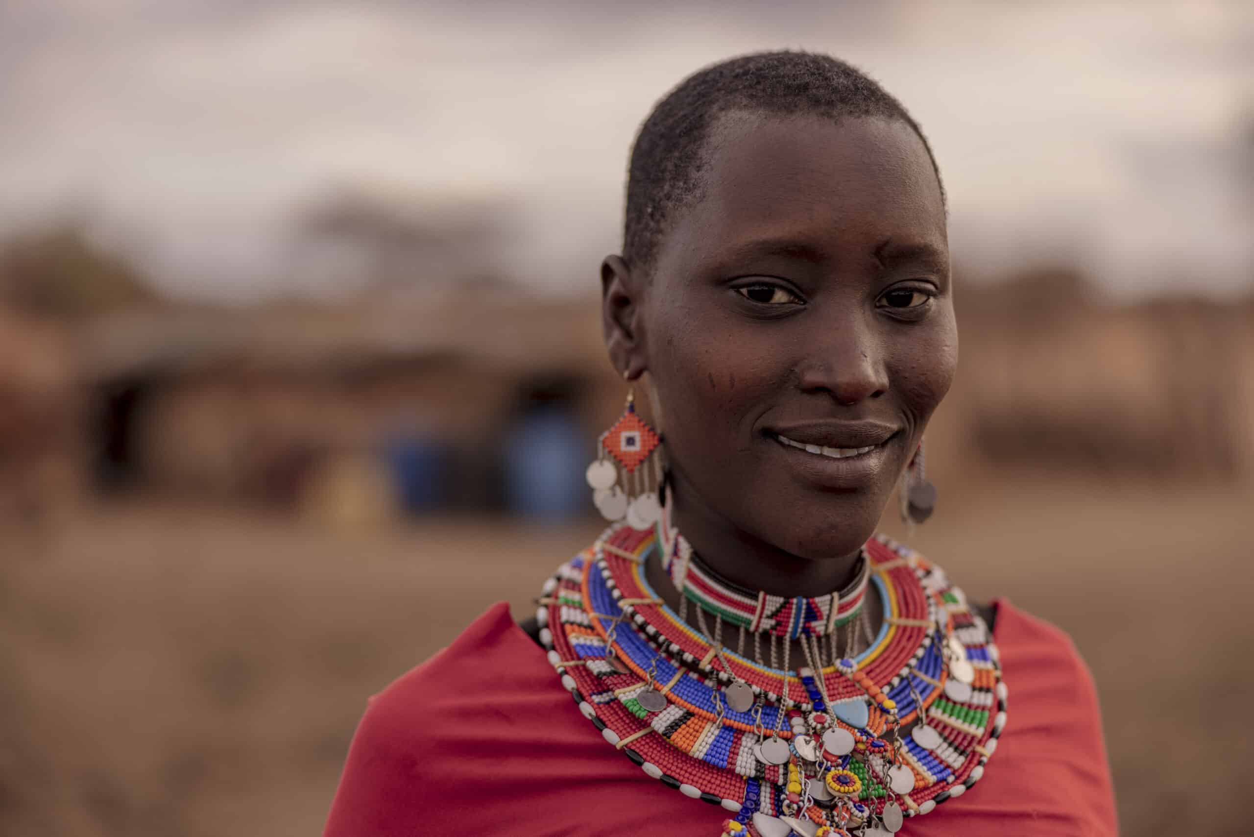 Ein Nahaufnahmeporträt einer lächelnden Frau in traditioneller Kleidung, einschließlich einer farbenfrohen Perlenkette und Ohrringen, vor dem unscharfen Hintergrund einer ländlichen Umgebung. © Fotografie Tomas Rodriguez