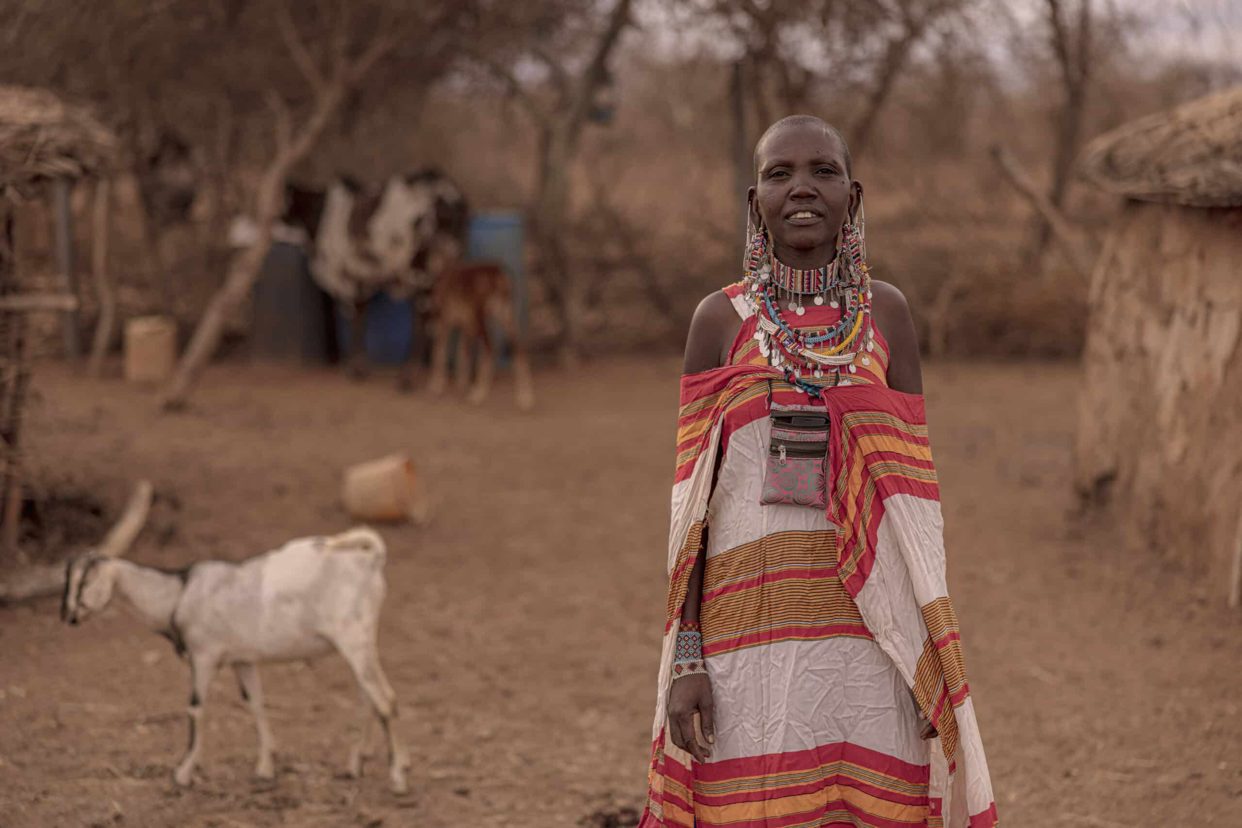 Eine Massai-Frau in traditioneller Kleidung steht in einem Dorf. Im Hintergrund sind Hütten und eine Ziege zu sehen, was eine heitere, ländliche Umgebung darstellt. © Fotografie Tomas Rodriguez