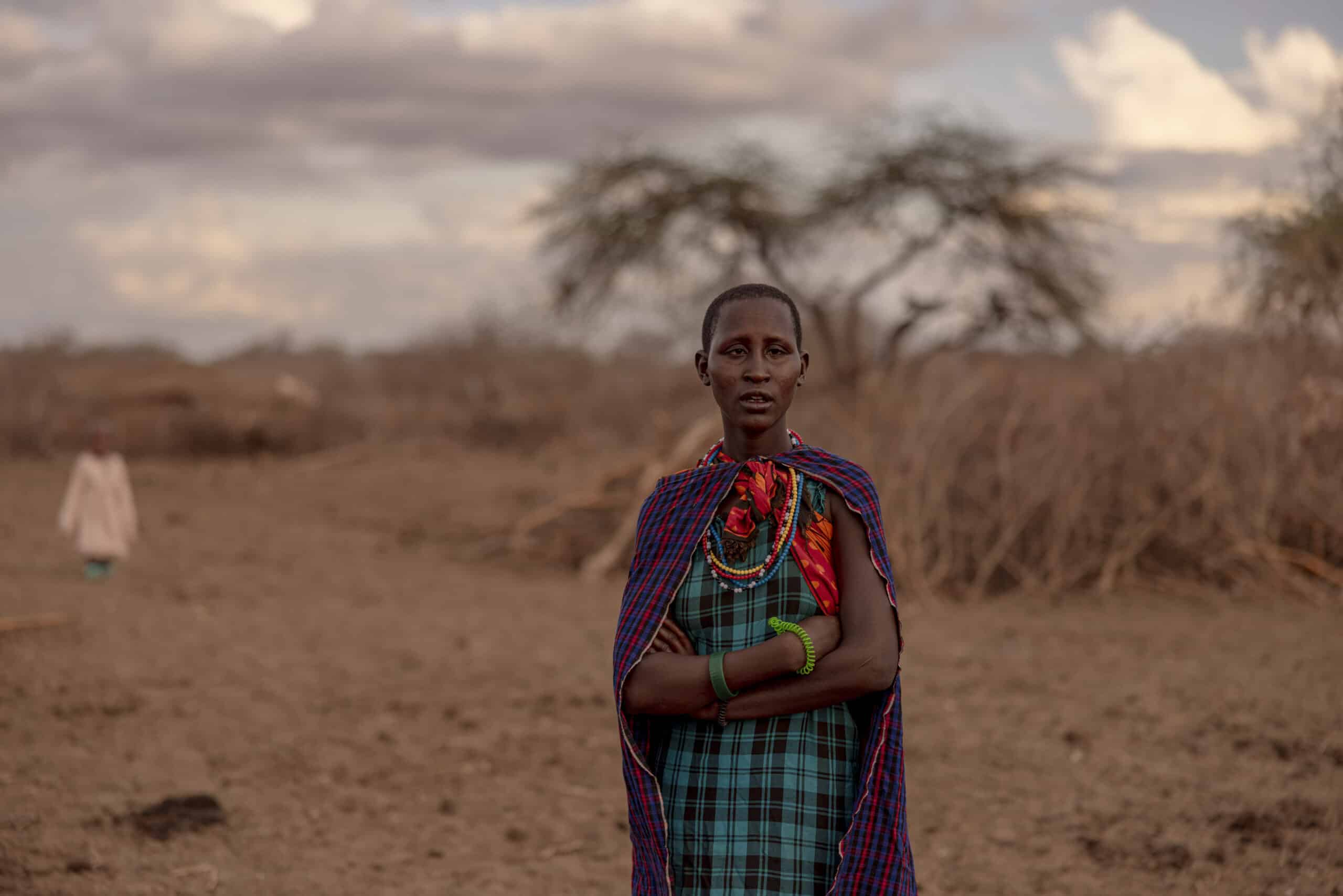 Eine Frau in traditioneller Massai-Kleidung steht mit verschränkten Armen in einer trockenen Landschaft, umgeben von spärlicher Vegetation unter einem bewölkten Himmel. © Fotografie Tomas Rodriguez