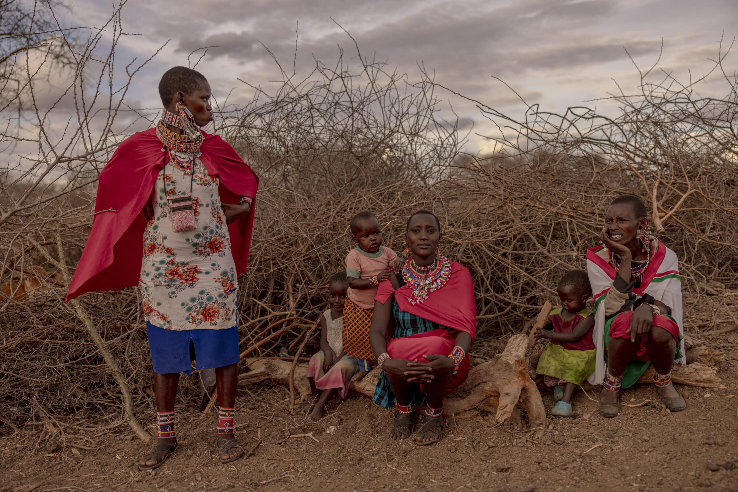 Ein Gruppenfoto einer Massai-Familie in ländlicher Umgebung: ein stehender Mann im roten Shuka telefoniert, drei sitzende Frauen, geschmückt mit bunten Perlen, zwei halten Kleinkinder und ein Mädchen vor der Kulisse eines Dornbusches. © Fotografie Tomas Rodriguez