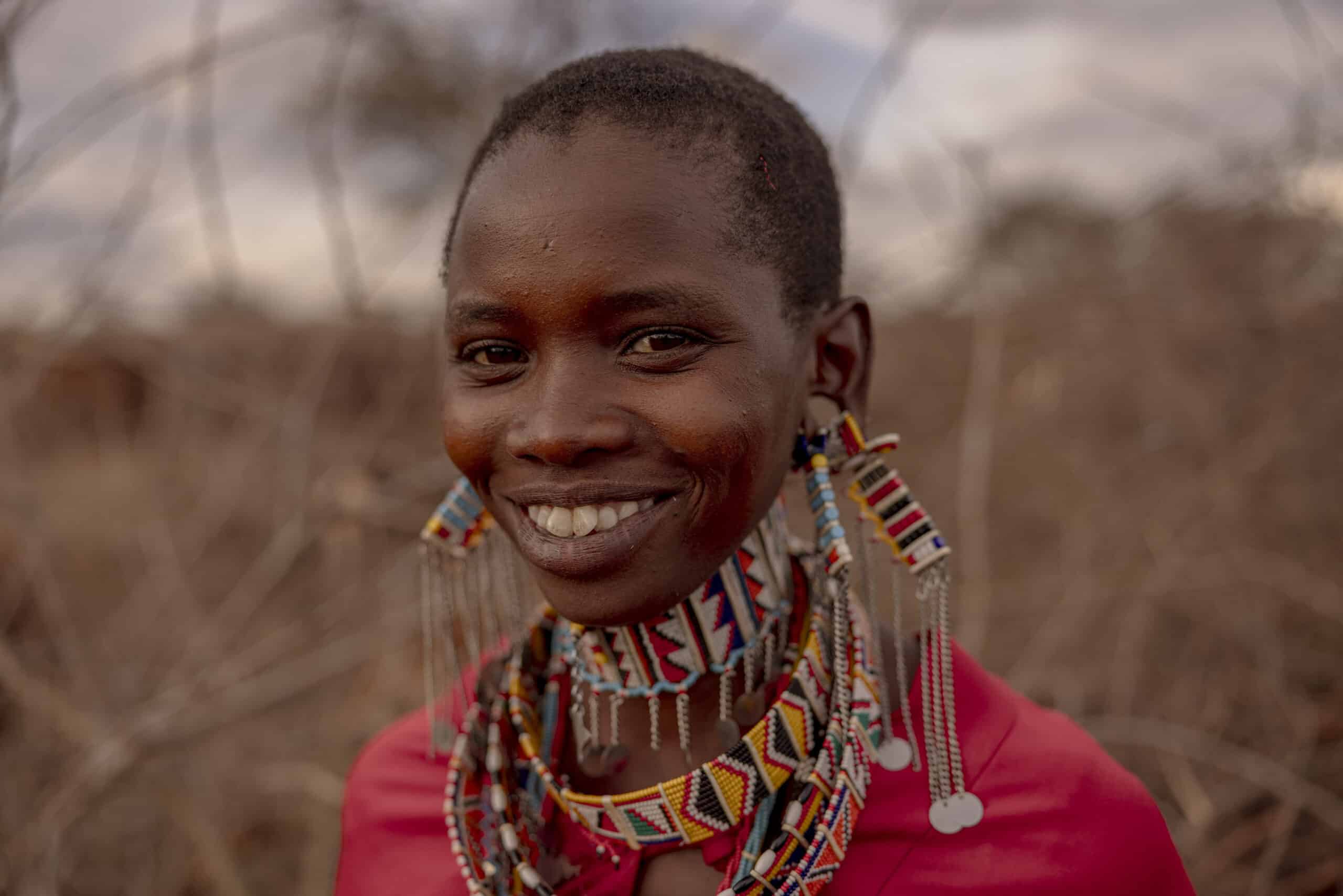 Eine lächelnde Frau mit kurzen Haaren, die aufwendigen Perlenschmuck und ein leuchtend rotes Oberteil trägt, vor einem unscharfen natürlichen Hintergrund. © Fotografie Tomas Rodriguez