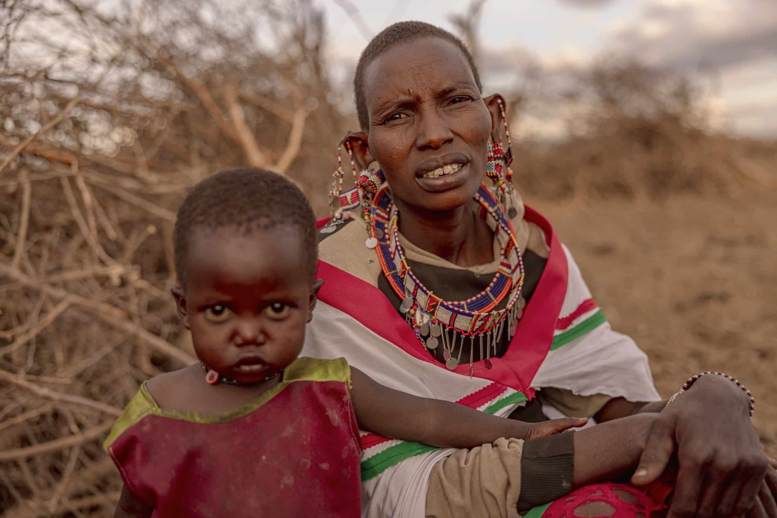 Eine Frau in traditioneller Massai-Kleidung und farbenfroher Perlenstickerei und ein kleines Kind in einem roten Gewand sitzen in der Abenddämmerung im Freien in einer trockenen Landschaft. Die Frau blickt in die Kamera, während das Kind ernst wirkt. © Fotografie Tomas Rodriguez