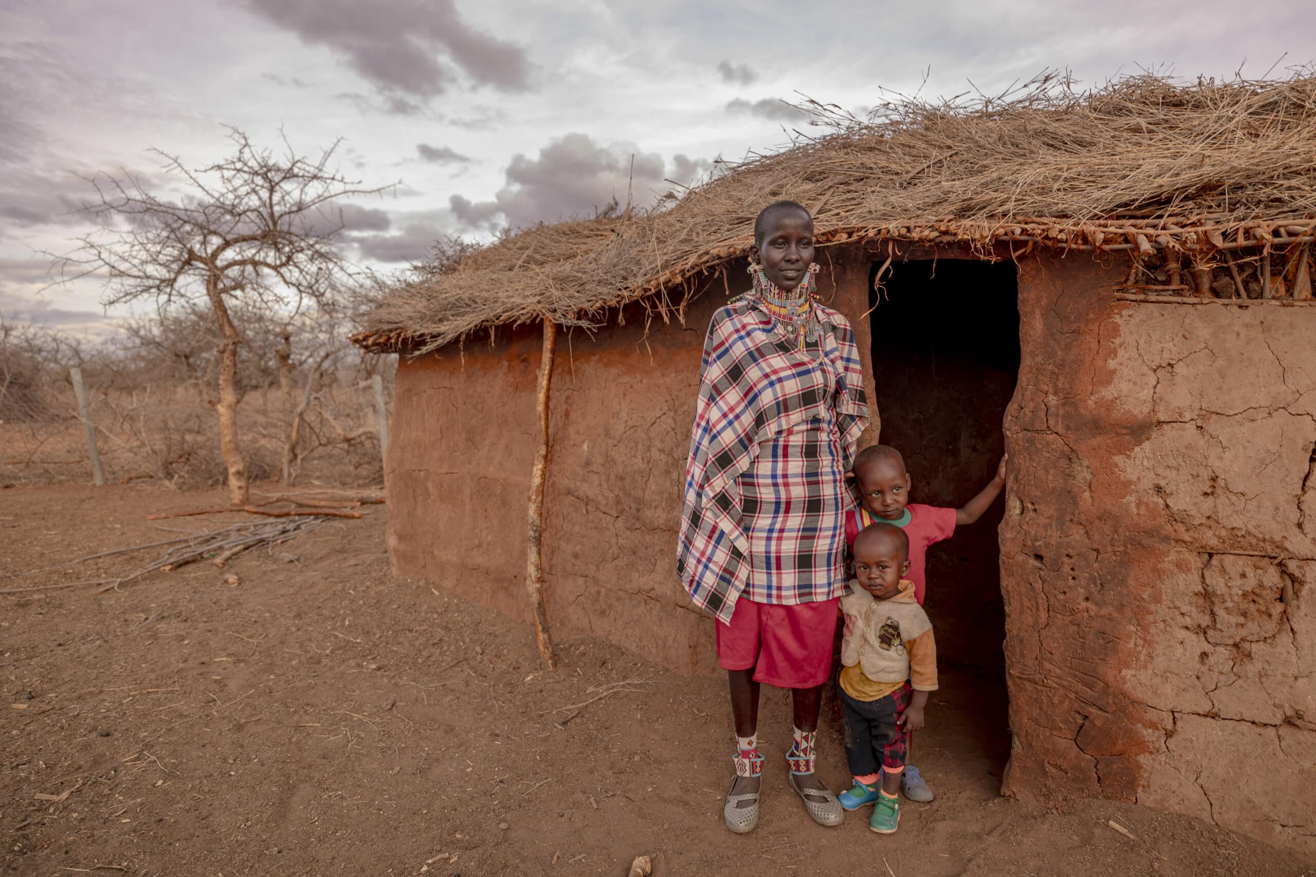 Eine Frau in traditioneller Kleidung steht mit zwei kleinen Kindern vor einer Lehmhütte mit Strohdach vor einer kargen Landschaft unter einem bewölkten Himmel. © Fotografie Tomas Rodriguez