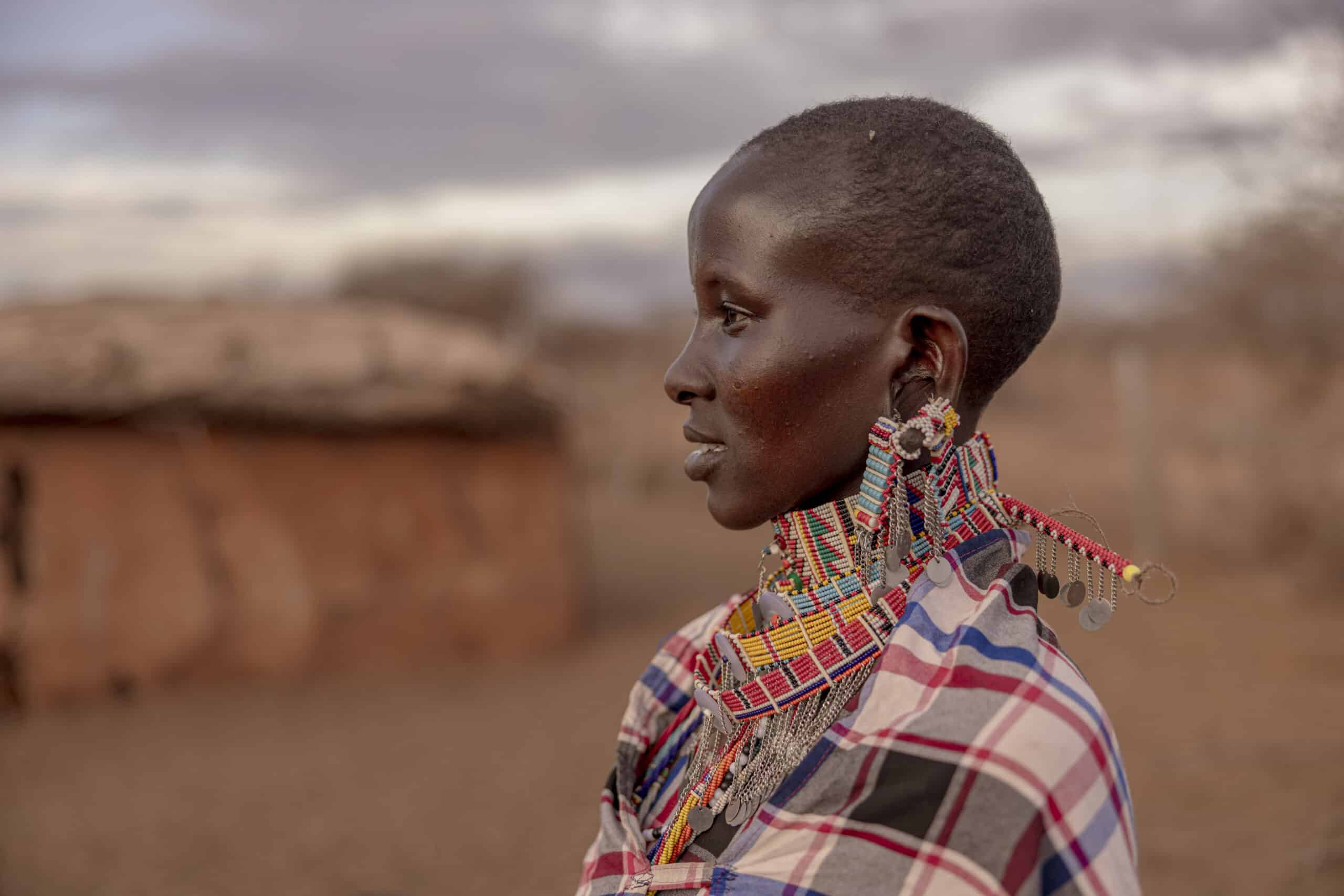 Das Profil einer jungen Frau vor einer ländlichen Kulisse. Sie trägt traditionellen Perlenschmuck und einen bunten Schal. Ihr Gesichtsausdruck ist nachdenklich. Im Hintergrund sind Hütten mit Strohdächern zu sehen. © Fotografie Tomas Rodriguez