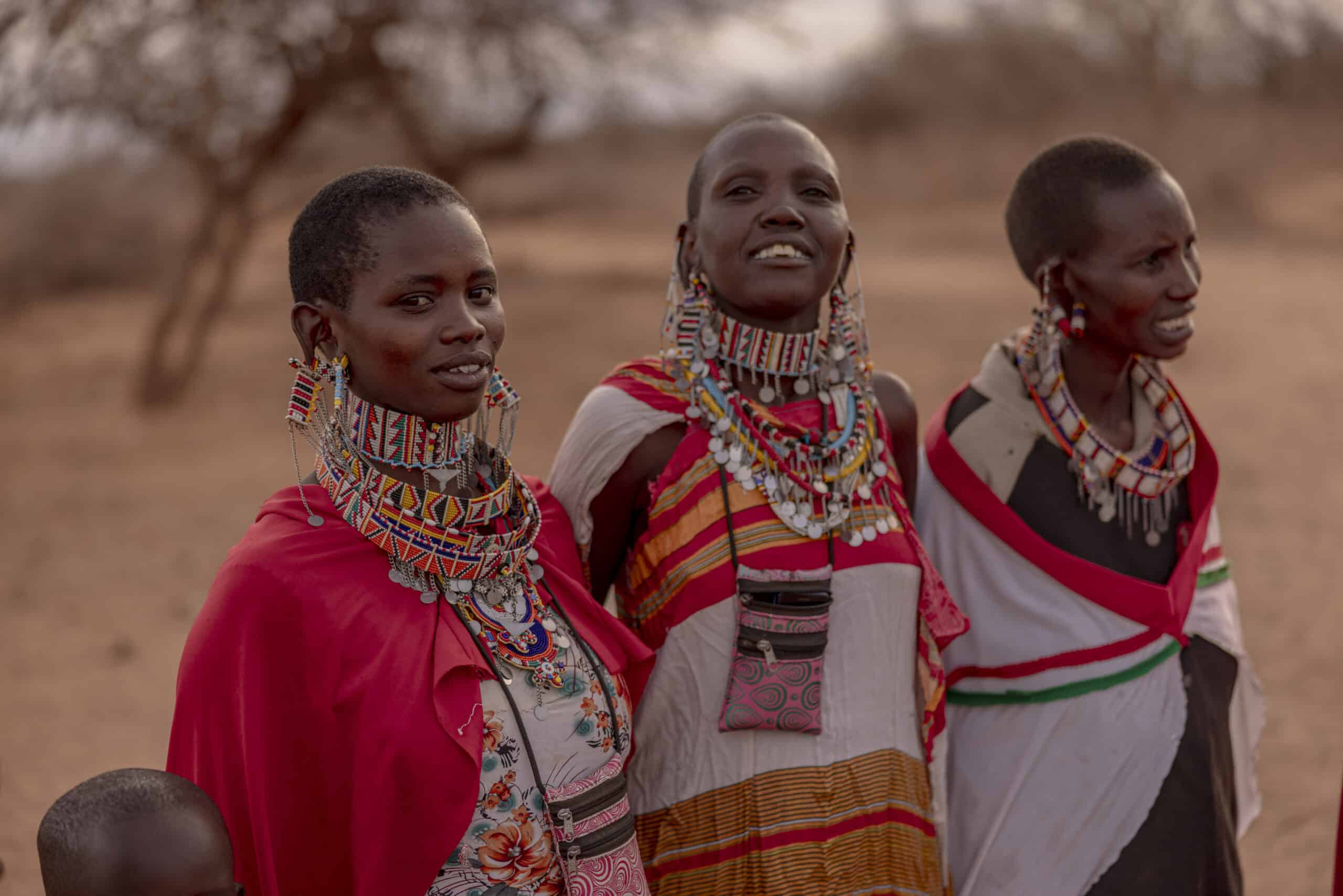 Drei Massai-Frauen, geschmückt mit bunten Halsketten und traditioneller Kleidung, stehen in einer Savannenlandschaft, lächeln und unterhalten sich miteinander. © Fotografie Tomas Rodriguez