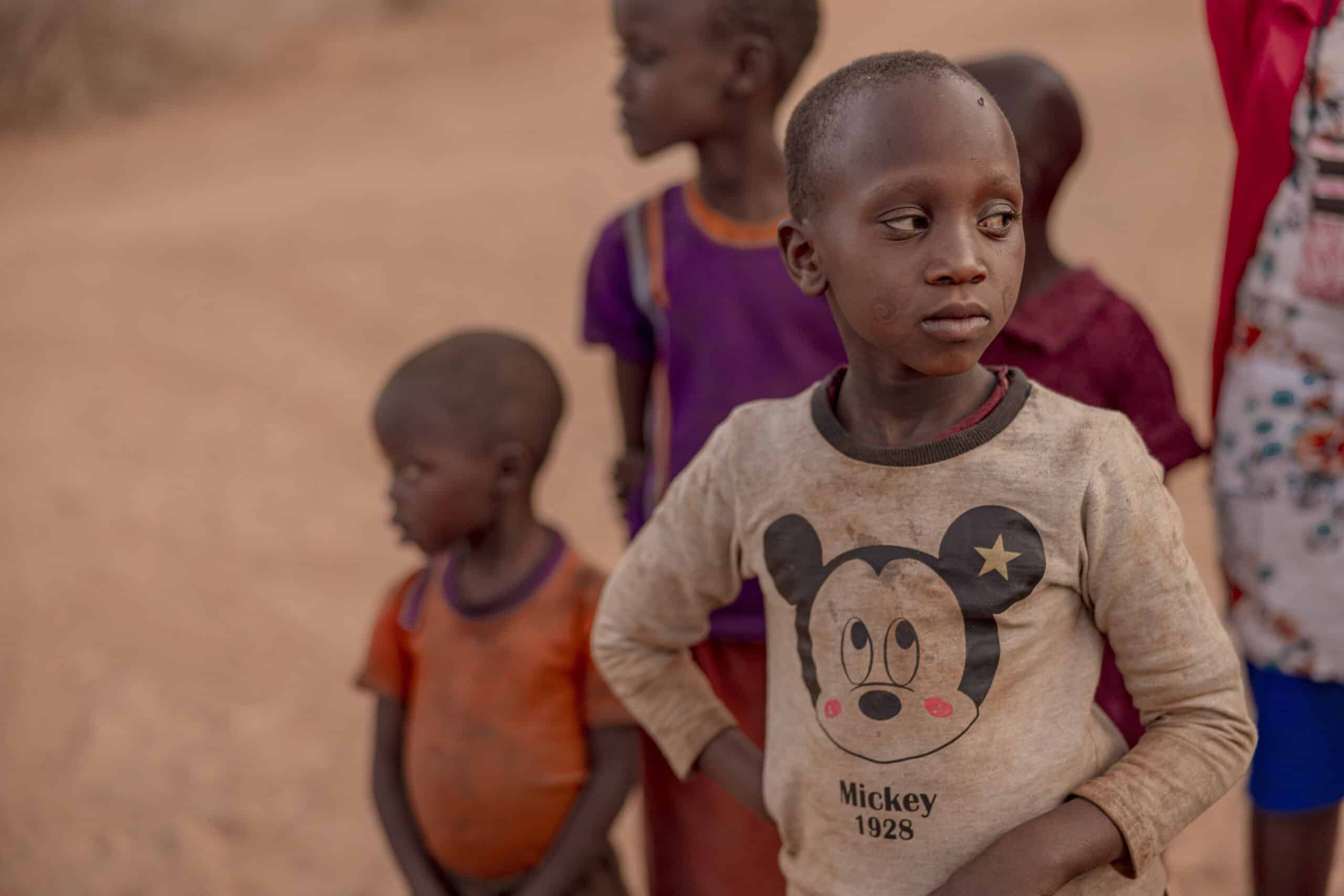 Eine Gruppe von Kindern mit einem Jungen im Vordergrund, der ein Mickey-Mouse-T-Shirt trägt und nachdenklich in einer staubigen Gegend steht. Um ihn herum sind andere Kinder zu sehen. © Fotografie Tomas Rodriguez