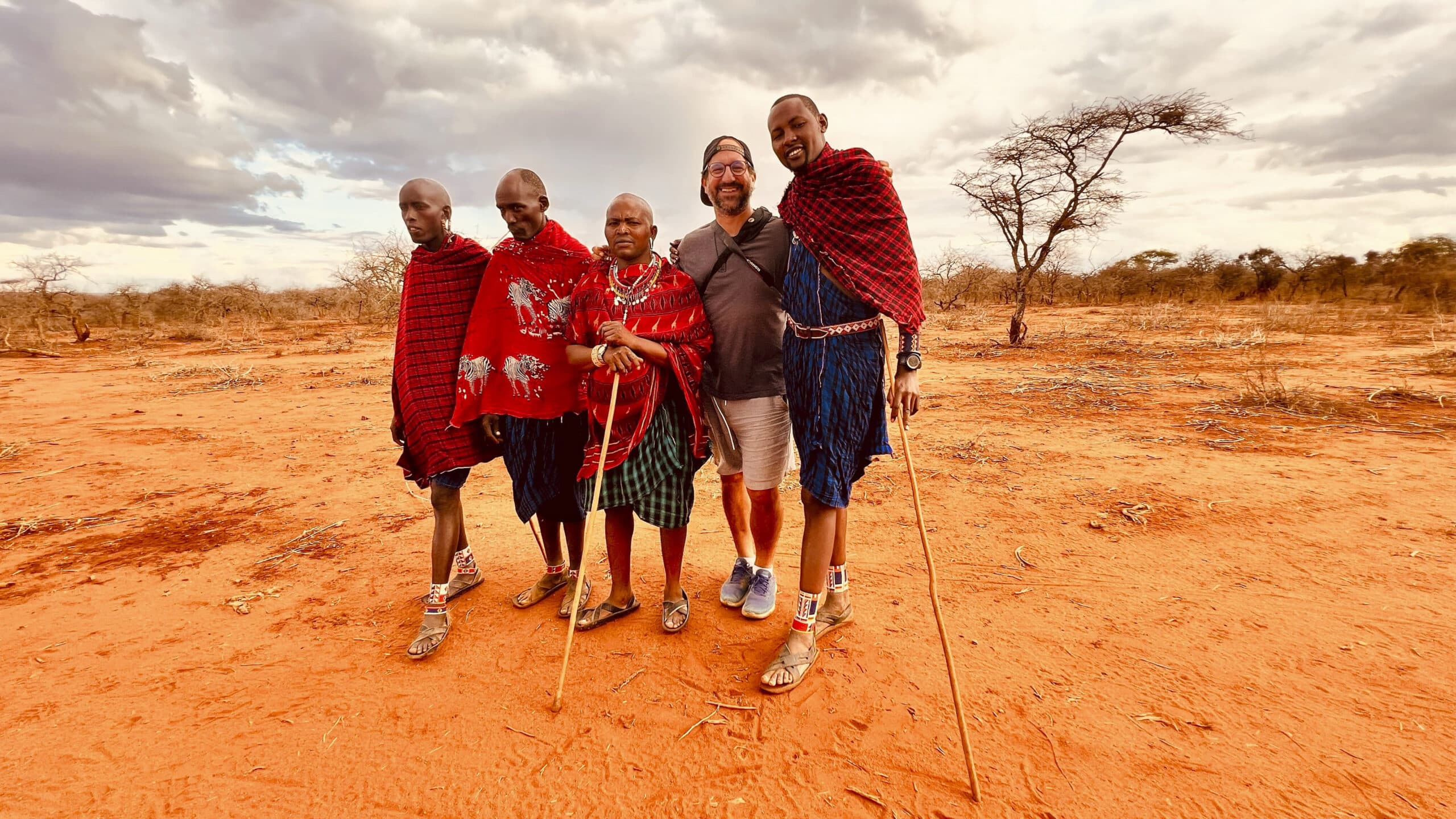 Fünf Menschen, darunter drei in traditioneller Massai-Kleidung, stehen auf einem roten Sandgelände mit spärlicher Vegetation und einem bewölkten Himmel im Hintergrund. © Fotografie Tomas Rodriguez