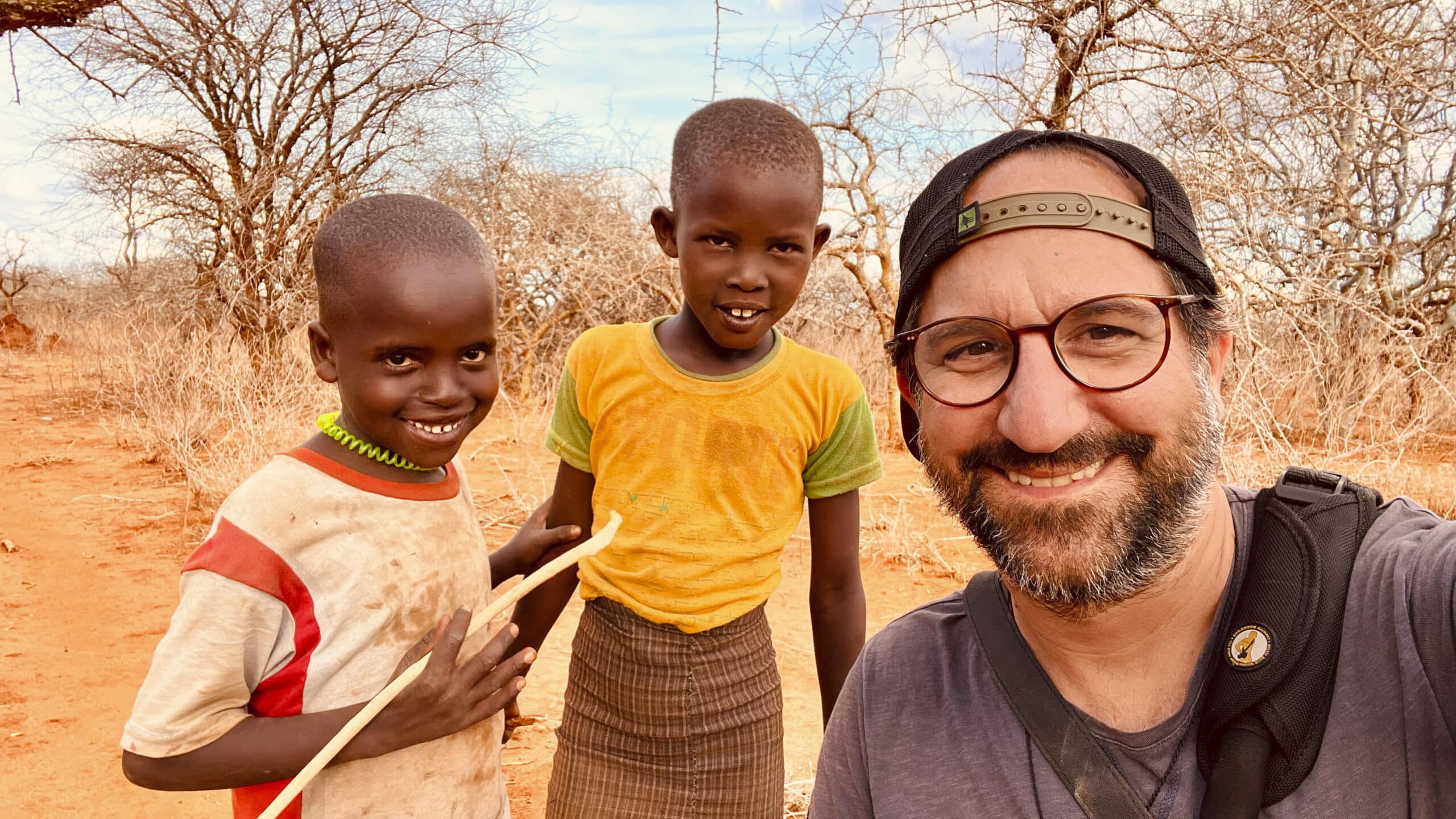 Ein Mann macht ein Selfie mit zwei kleinen Kindern in einer trockenen, buschigen Landschaft. Die Kinder lächeln und eines hält einen Stock, während im Hintergrund dornige Büsche und kahle Bäume zu sehen sind. © Fotografie Tomas Rodriguez