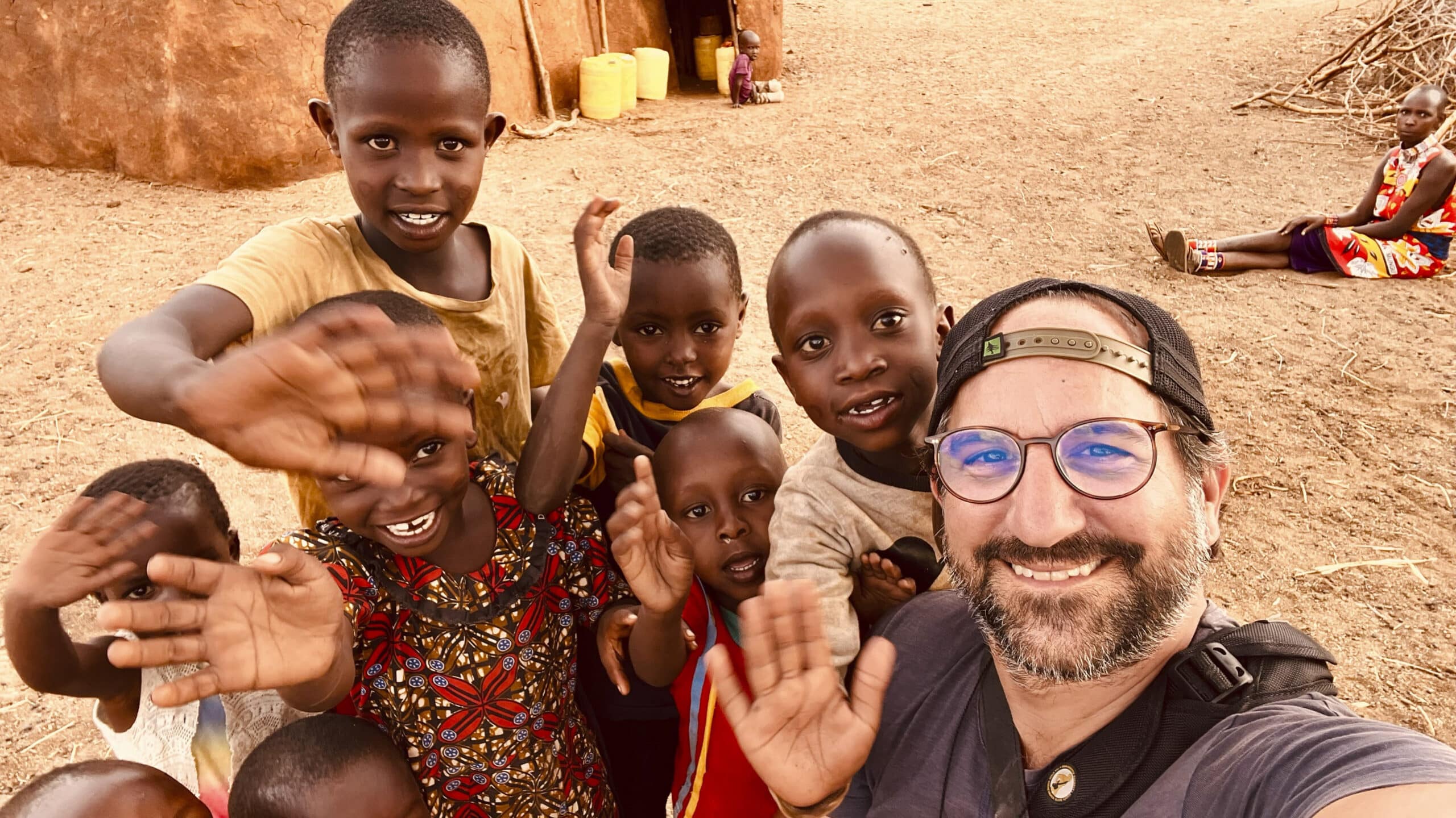 Ein fröhliches Selfie eines Mannes mit einer Gruppe von sechs lächelnden Kindern in einer ländlichen Umgebung, die in die Kamera winken. Im Hintergrund sind traditionelle Hütten und eine sitzende Frau zu sehen. © Fotografie Tomas Rodriguez