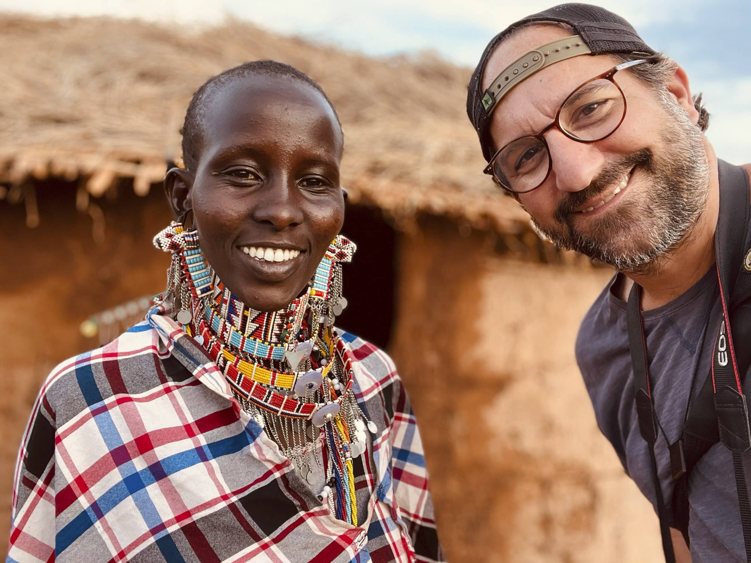 Eine lächelnde, mit bunten Perlenketten geschmückte Massai-Frau steht neben einem Mann mit Mütze und Brille vor einer Strohhütte. © Fotografie Tomas Rodriguez