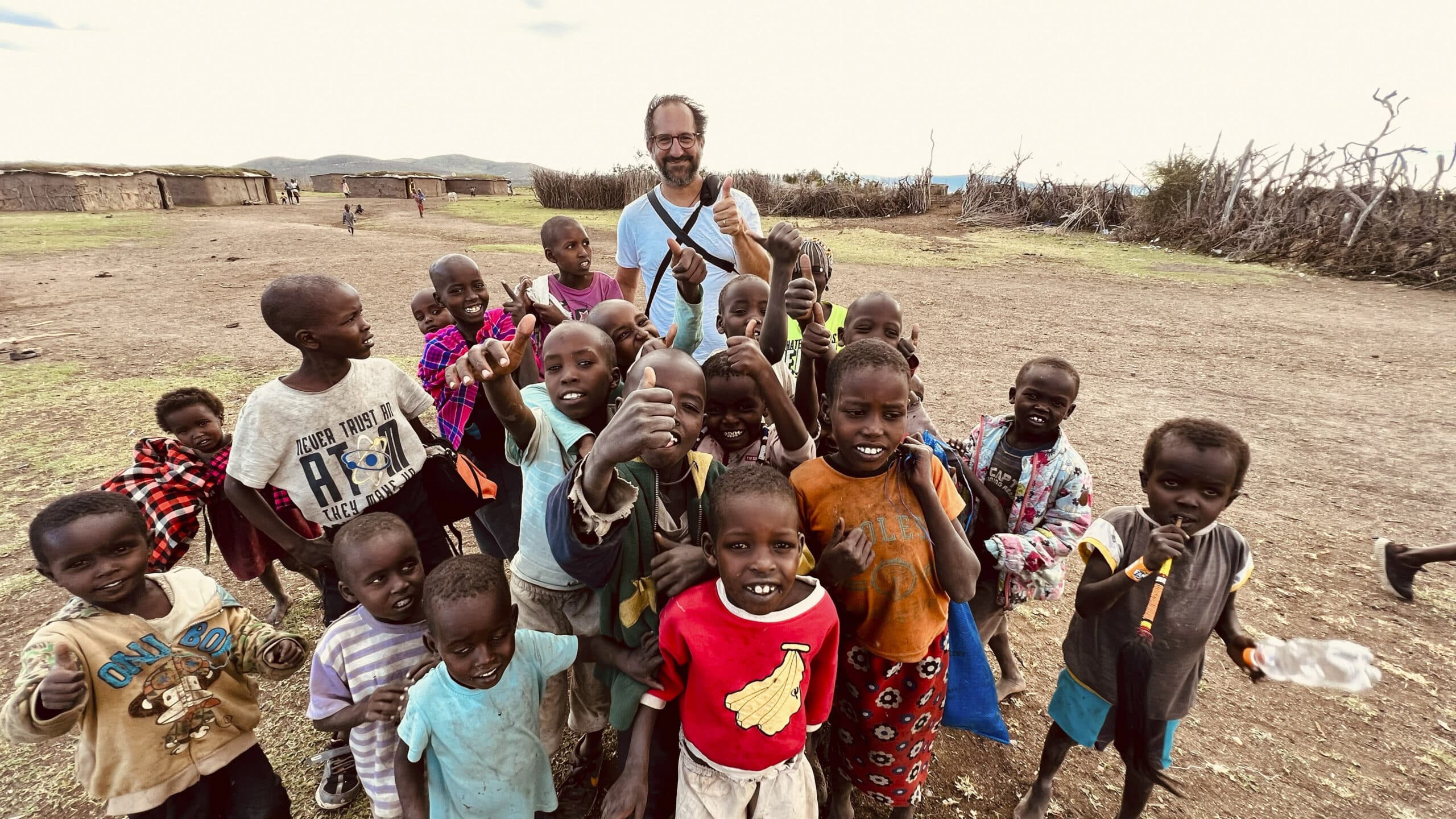 Ein Mann steht umgeben von einer Gruppe fröhlicher Kinder in einer ländlichen, trockenen Landschaft. Sie alle lächeln in die Kamera, einige winken, im Hintergrund sind offene Felder und ein paar Bäume zu sehen. © Fotografie Tomas Rodriguez