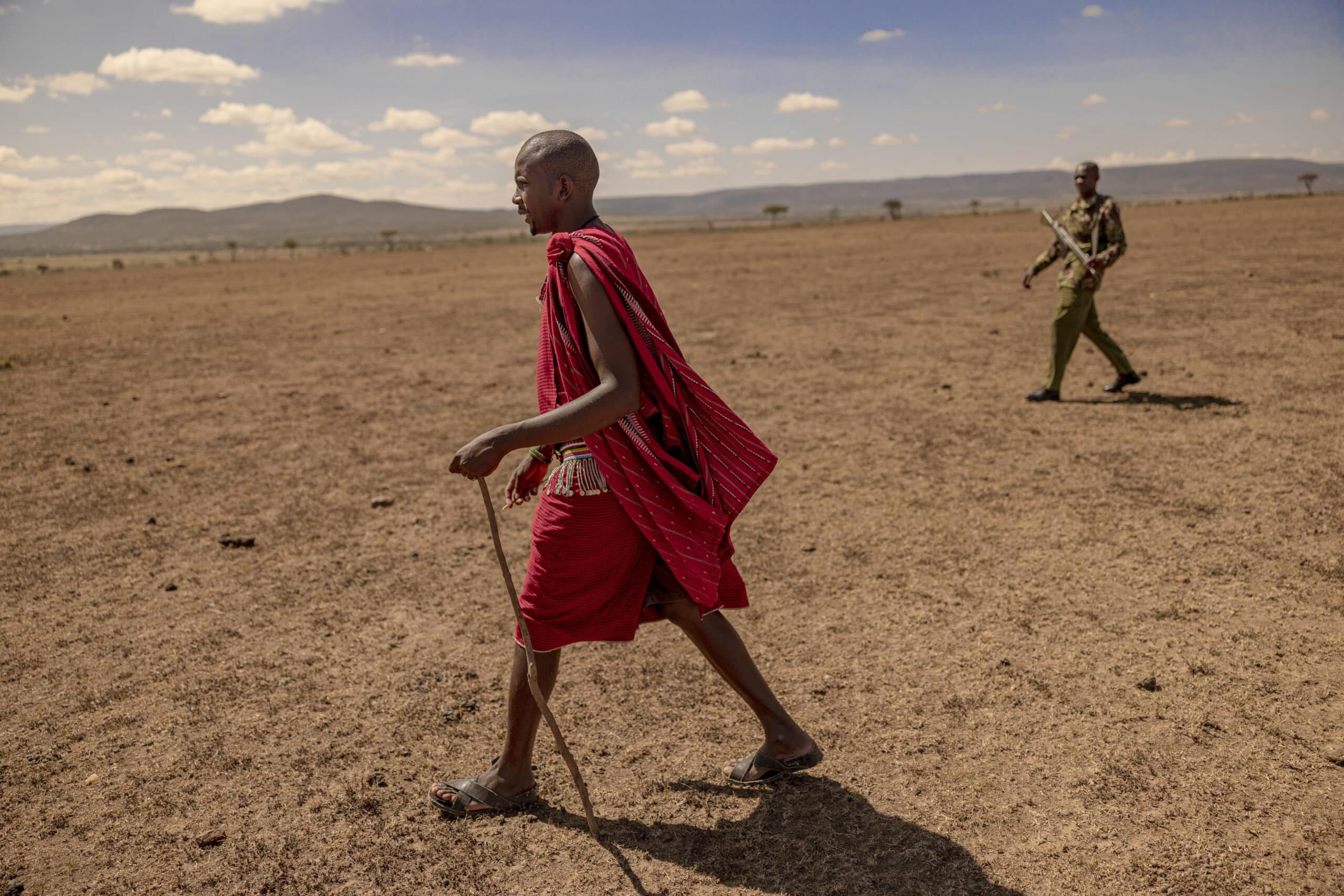 Ein Massai-Mann in traditioneller roter Kleidung geht durch ein trockenes Grasland. Im Hintergrund ist unter einem weiten, wolkigen Himmel ein weiterer Mann zu sehen. © Fotografie Tomas Rodriguez