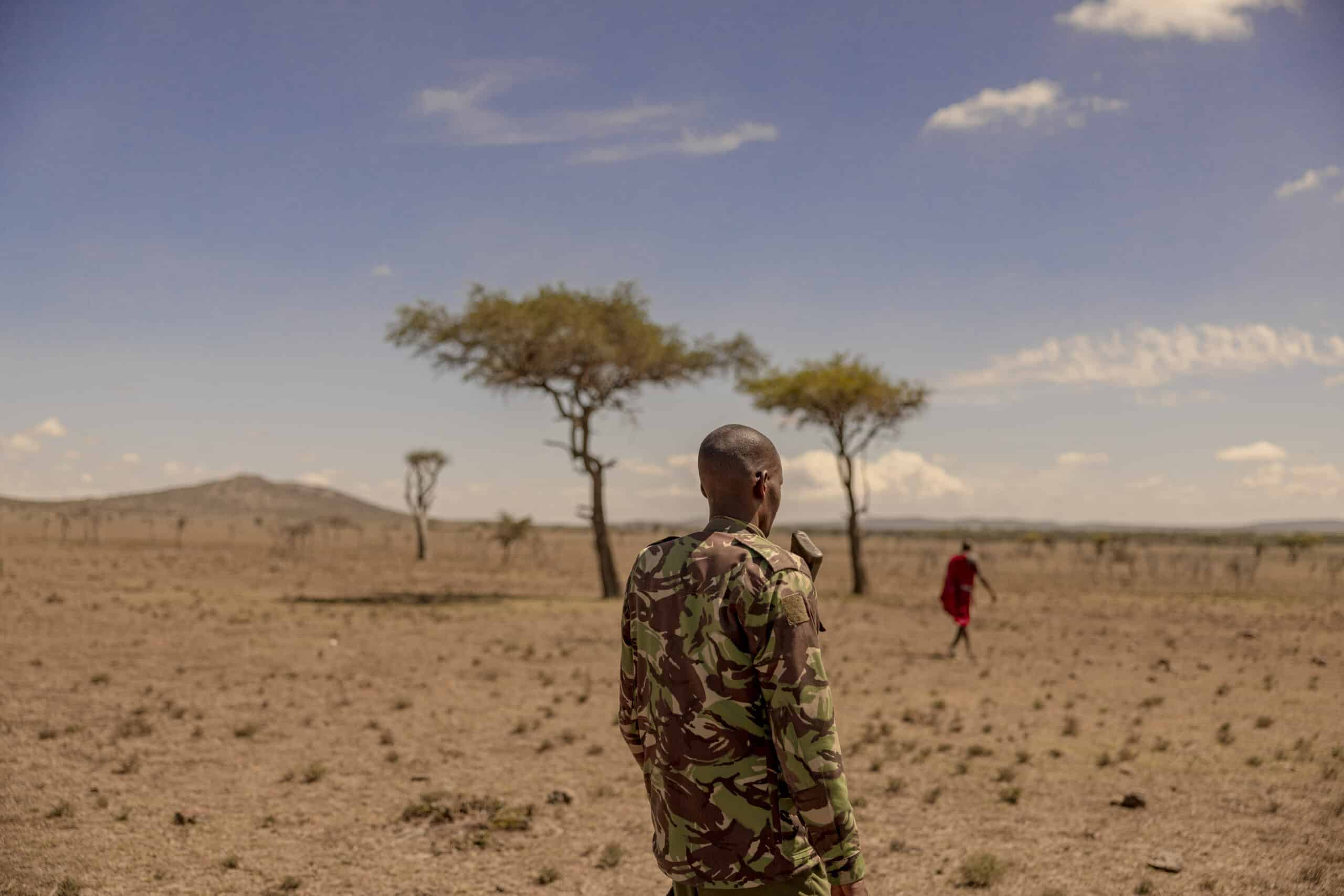 Eine Person in Militärtarnung steht mit dem Rücken zur Wand und blickt auf eine weite, trockene Landschaft mit vereinzelten Bäumen unter einem bewölkten Himmel. In der Ferne geht eine weitere Person in Rot davon. © Fotografie Tomas Rodriguez