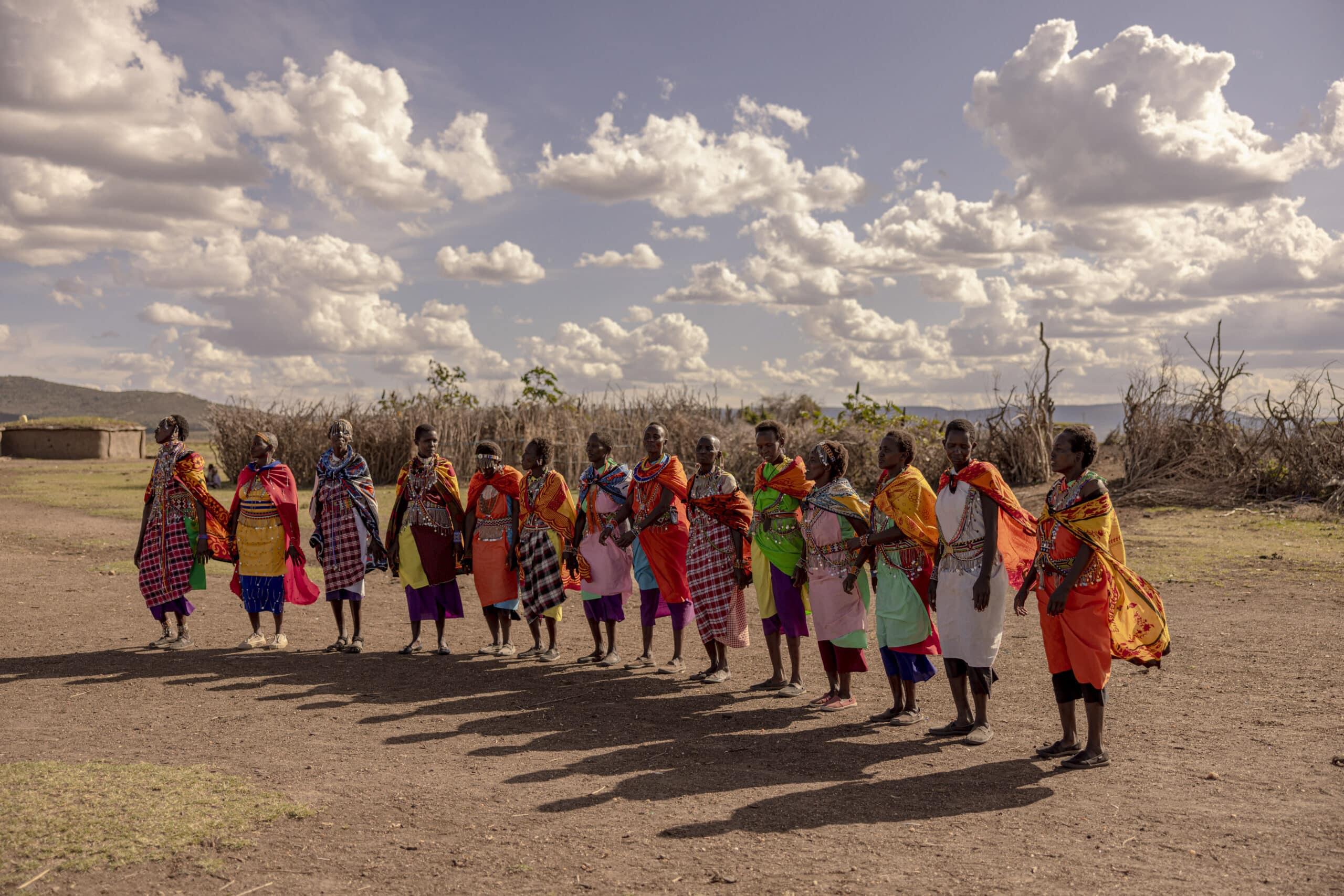 Eine Gruppe von Massai-Frauen in traditioneller, farbenfroher Kleidung steht in einer Reihe in einer staubigen Savanne unter einem bewölkten Himmel, hält Händchen und scheint mitten im Tanz oder einer Feier zu sein. © Fotografie Tomas Rodriguez