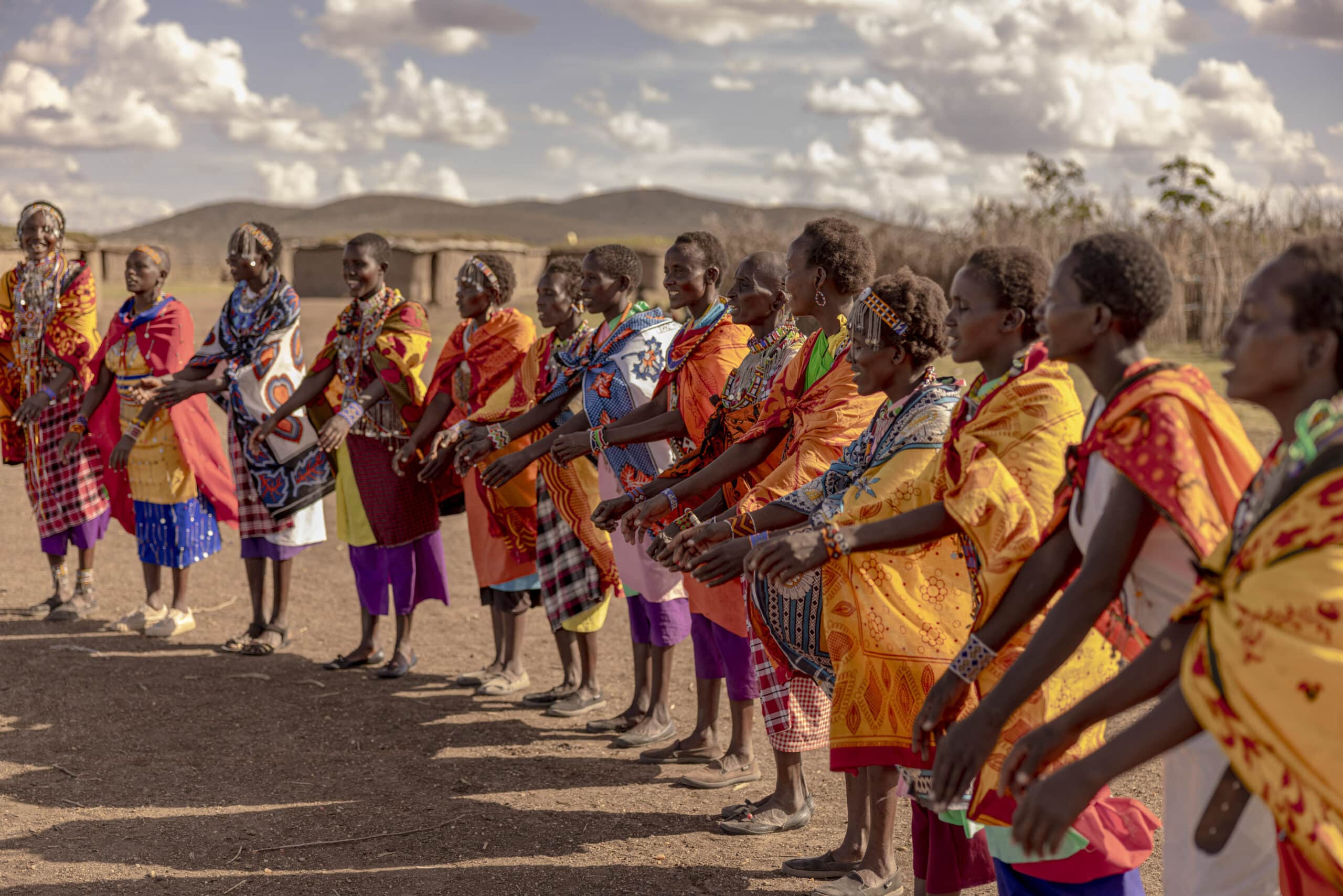 Eine Gruppe von Massai-Frauen in traditioneller heller Kleidung und Perlenschmuck steht im Freien in einer Reihe und nimmt unter klarem Himmel an einer kulturellen Darbietung teil. © Fotografie Tomas Rodriguez