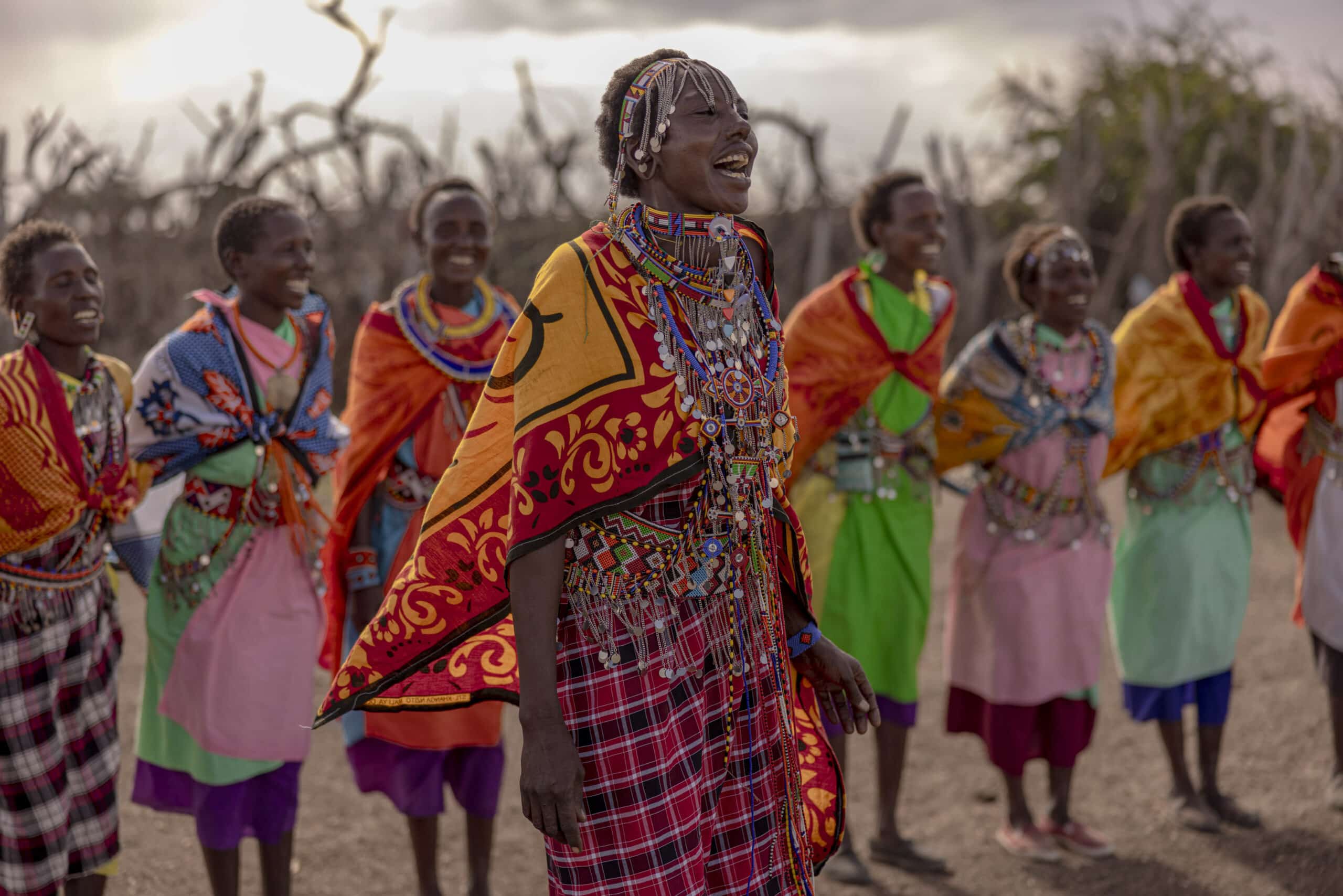 Eine Gruppe Massai-Frauen in traditioneller Kleidung, geschmückt mit bunten Perlen und Stoffen, tanzt fröhlich in der Abenddämmerung in einer trockenen, buschigen Landschaft. © Fotografie Tomas Rodriguez