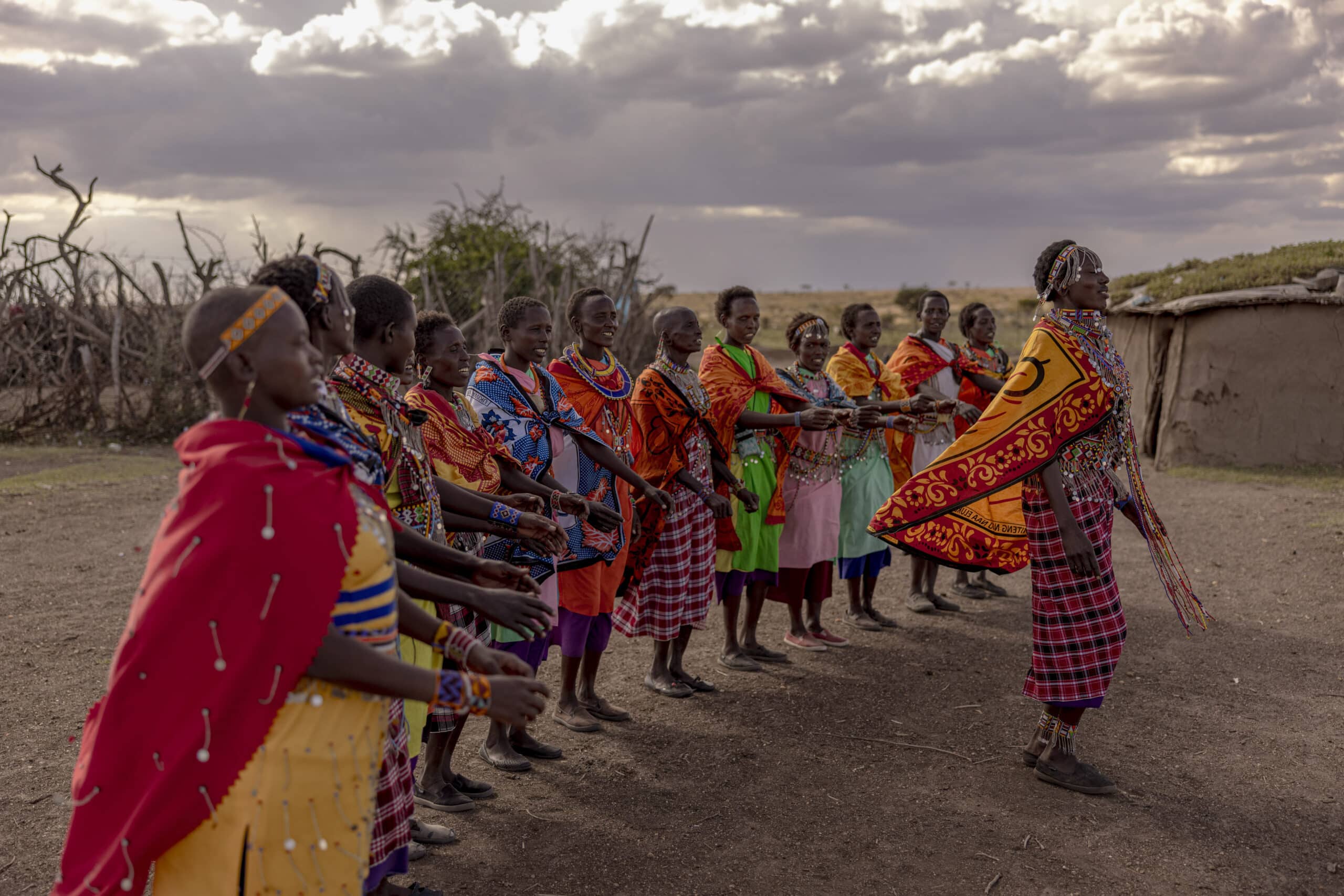 Eine Gruppe von Massai-Frauen in traditioneller, farbenfroher Kleidung führt in ländlicher Umgebung unter einem bewölkten Himmel einen kulturellen Tanz auf. © Fotografie Tomas Rodriguez