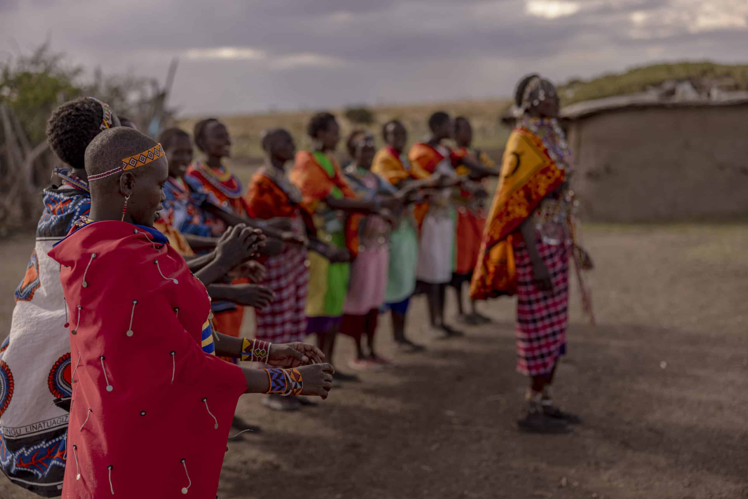 Eine Gruppe von Massai in traditioneller Kleidung nimmt an einer Gemeinschaftsversammlung oder Zeremonie teil. Im Vordergrund steht eine Frau in einem roten Shuka. © Fotografie Tomas Rodriguez