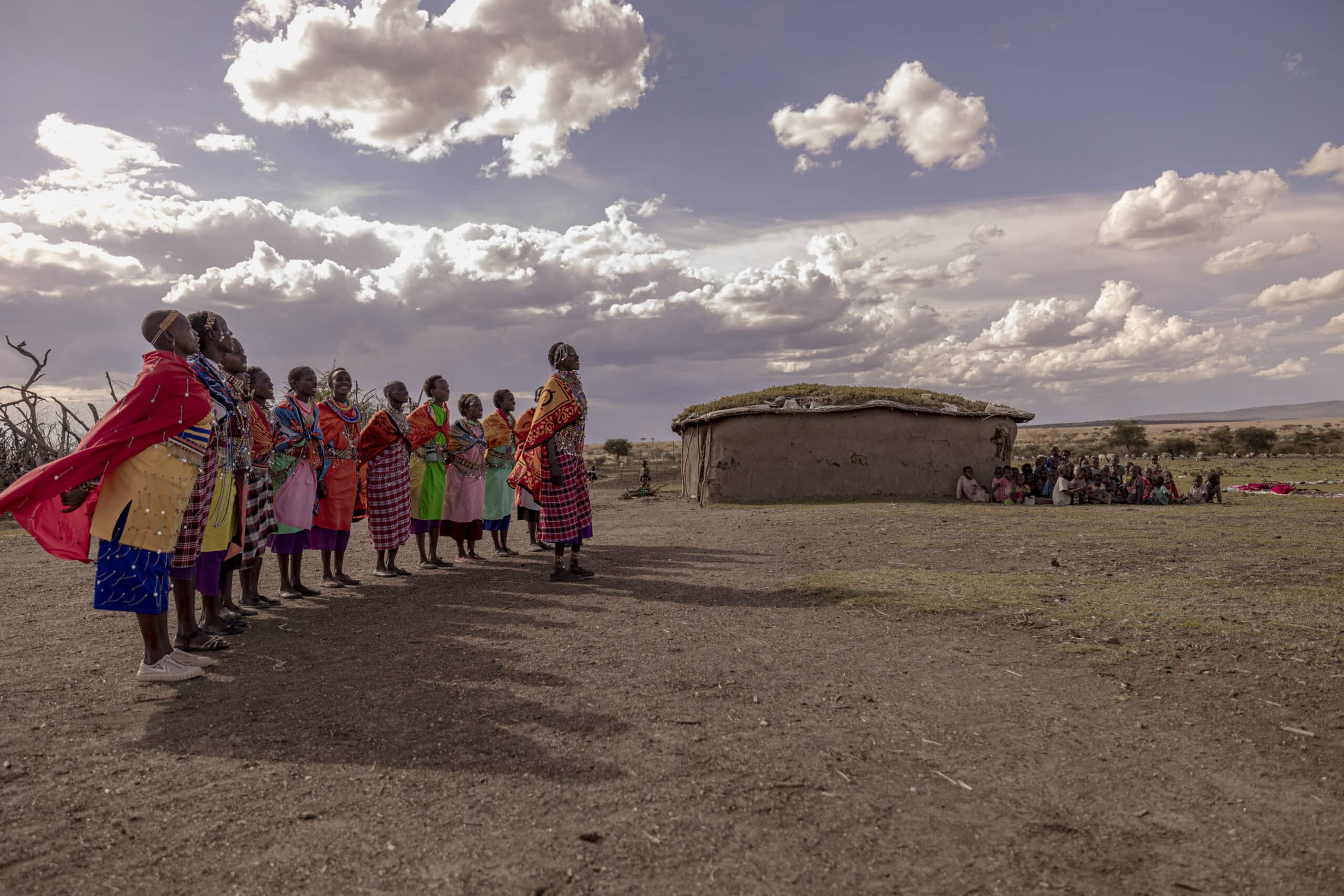 Eine Gruppe von Massai in traditioneller Kleidung steht in einer Reihe und führt unter einem weiten Himmel in der Nähe einer Lehmhütte einen Tanz auf. Im Hintergrund haben sich an diesem bewölkten Tag noch weitere Menschen versammelt. © Fotografie Tomas Rodriguez