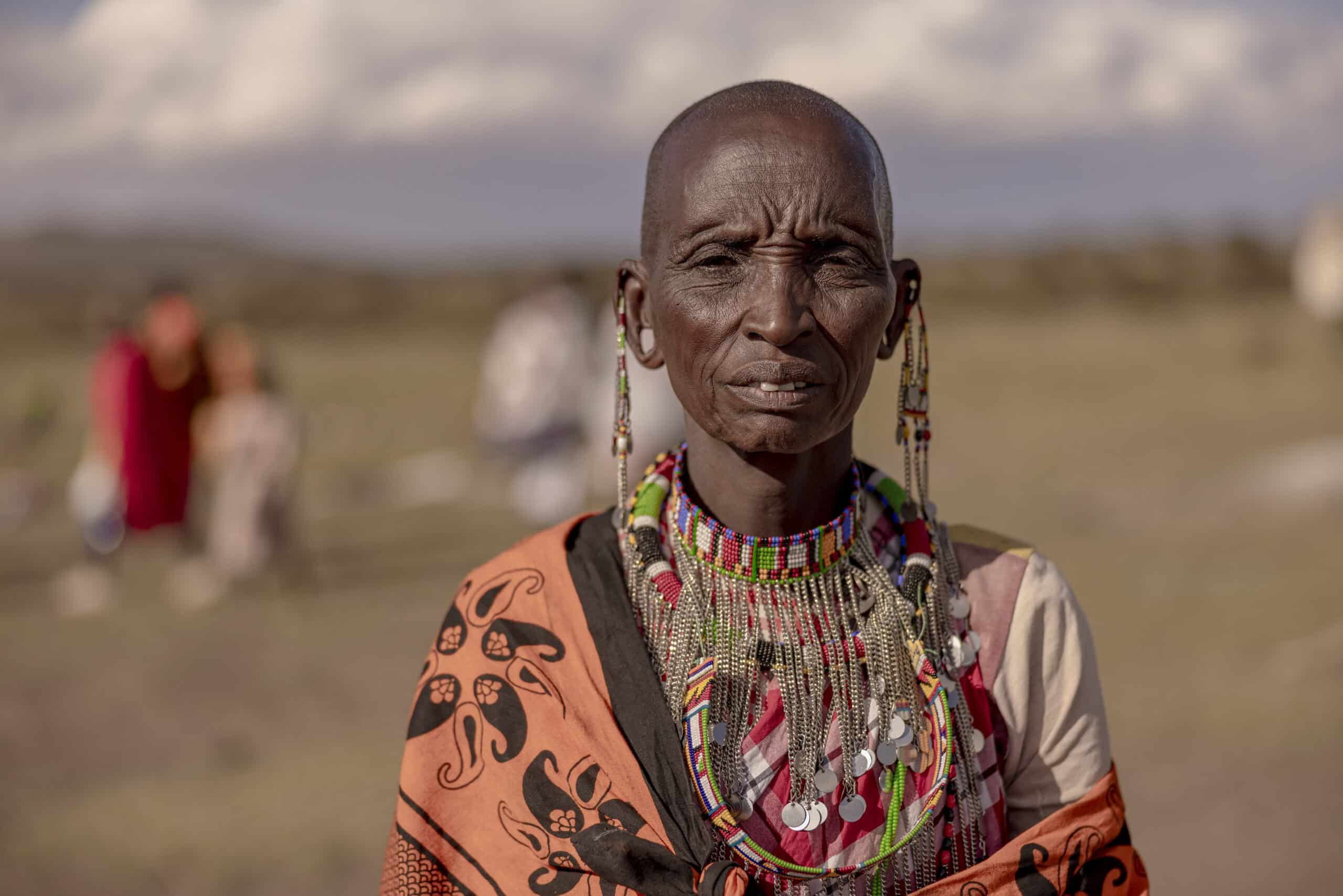 Eine Massai-Frau in traditioneller Kleidung, darunter Perlenschmuck und eine gemusterte Shuka, steht auf einem Feld, im Hintergrund sind verschwommene Figuren zu sehen. © Fotografie Tomas Rodriguez