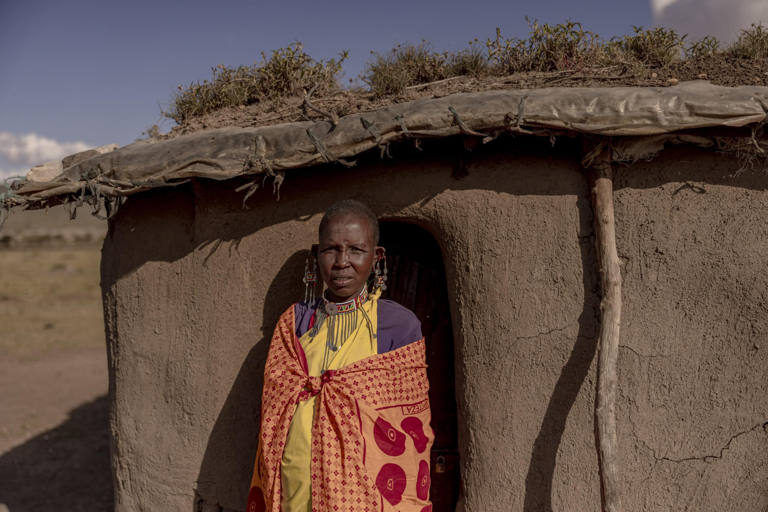 Eine Massai-Frau in traditioneller Kleidung steht vor einem Lehmhaus mit Grasdach in der Savanne. © Fotografie Tomas Rodriguez