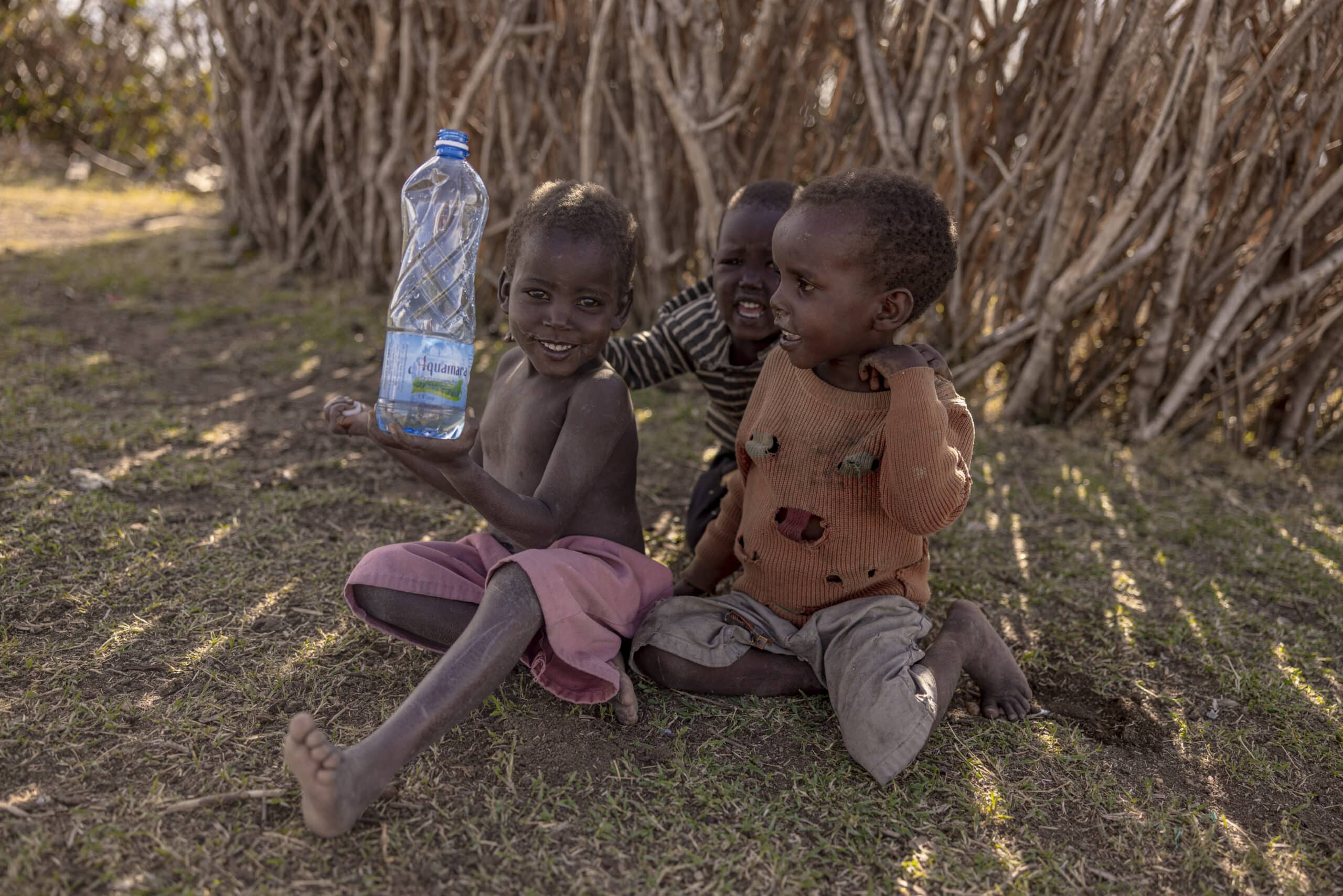 Drei kleine Kinder sitzen auf dem Boden neben einem Strohzaun. Ein Kind hält eine große Wasserflasche und lächelt, während die anderen fröhlich zuschauen. Sie befinden sich in einer ländlichen Umgebung mit natürlichem Licht. © Fotografie Tomas Rodriguez