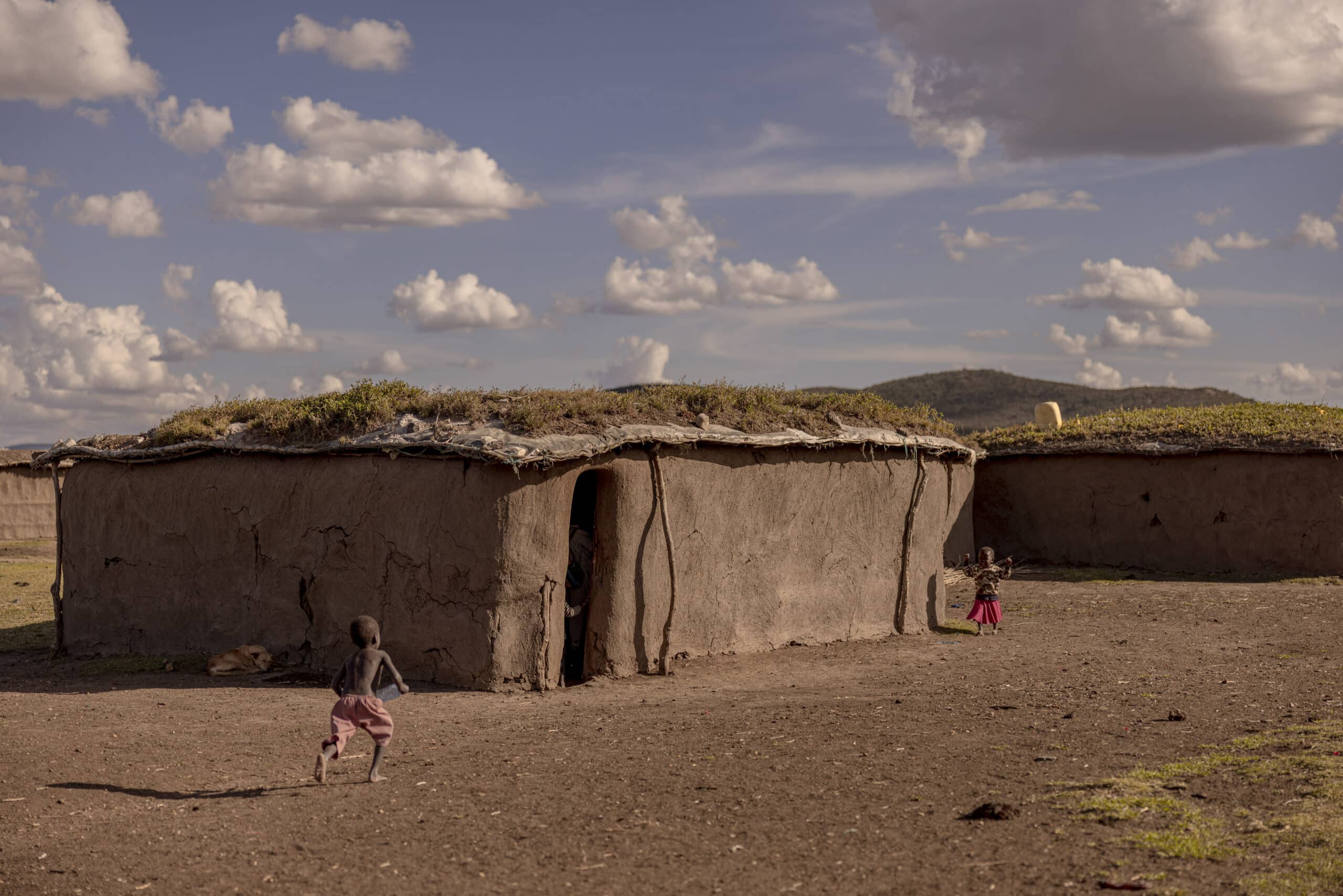 Zwei Kinder spielen neben traditionellen Lehmhütten mit Strohdächern in einer ländlichen Umgebung mit sanften Hügeln und einem bewölkten Himmel im Hintergrund. © Fotografie Tomas Rodriguez